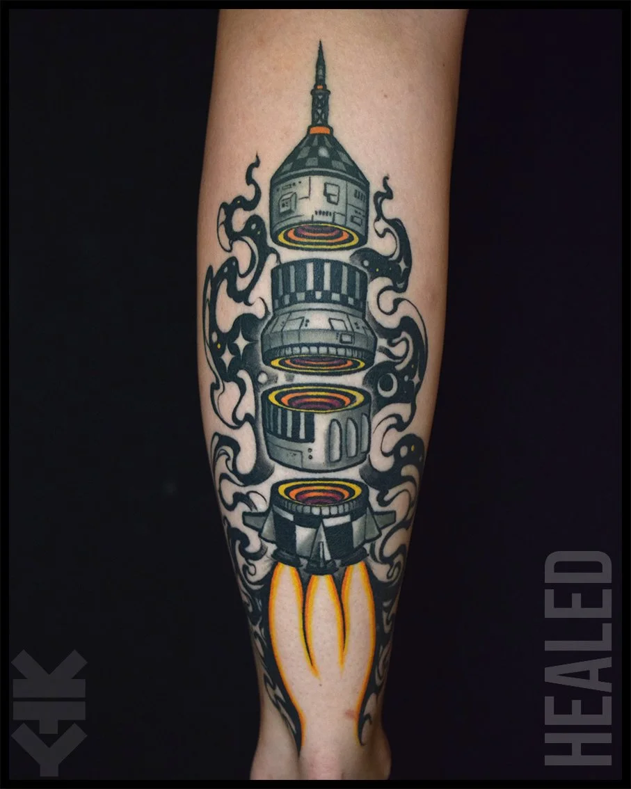 Tattoo of a rocket with a sci-fi design shooting fire from its base, surrounded by stylized black smoke.