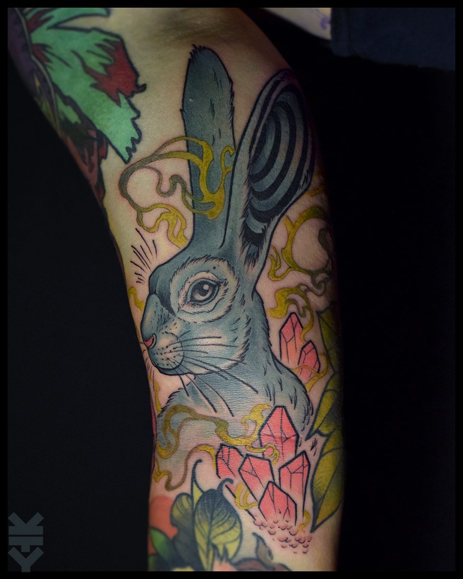 Tattoo of a bunny with large ears, detailed fur, and a whimsical background with pink crystals and yellow swirling lines.