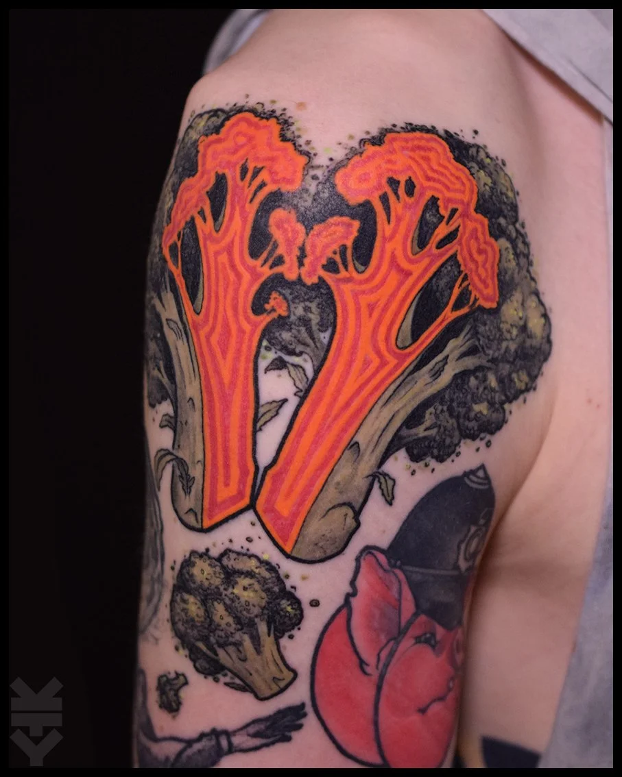 Tattoo of a large steaming bowl of ramen with orange broth and noodles, accompanied by broccoli and a raw egg on the person's upper arm.