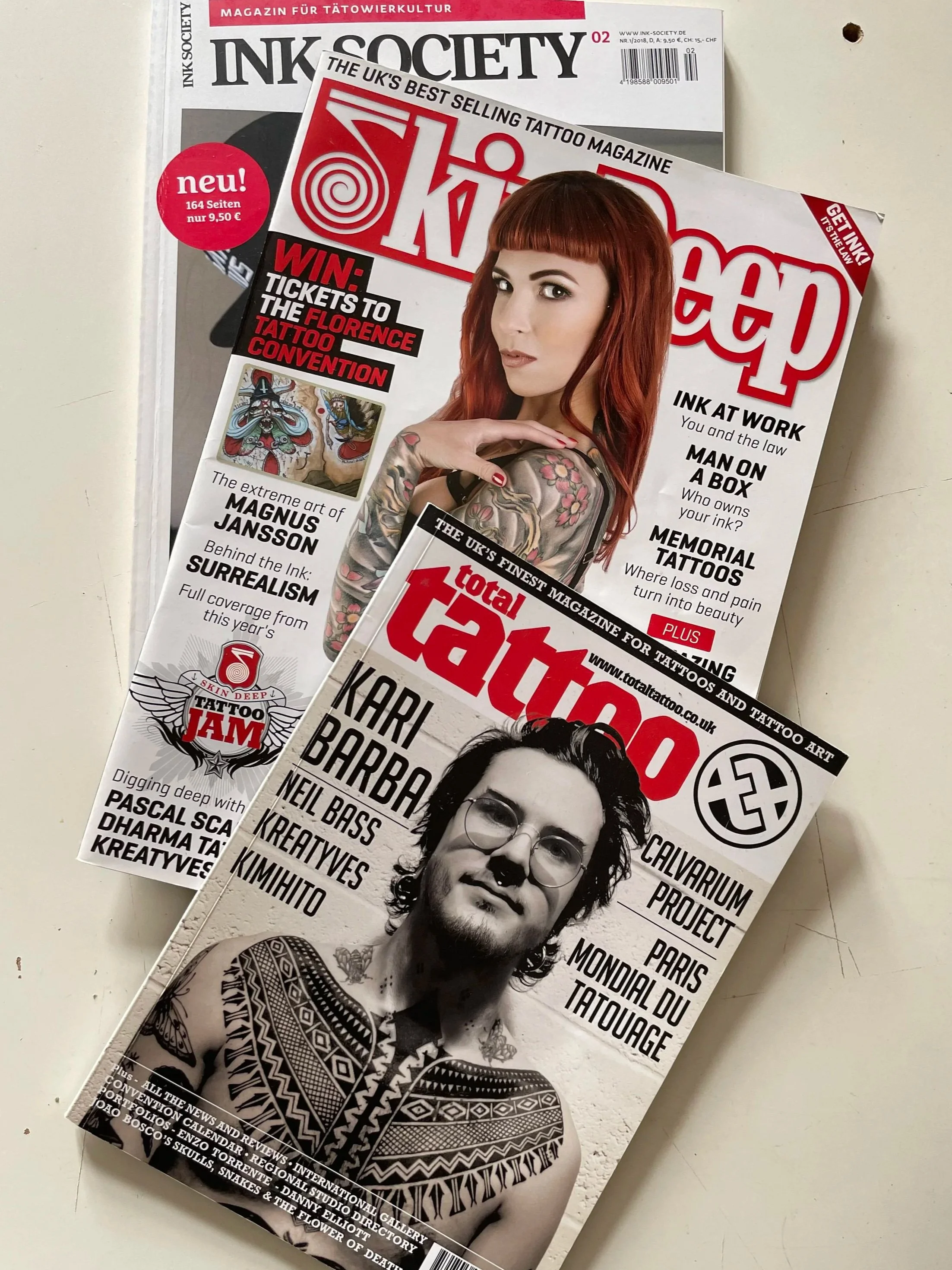 Two tattoo magazines stacked on a white surface. The top magazine features a woman with red hair and tattoos on her arm, while the bottom magazine shows a man with tattoos, glasses, and a tribal tattoo on his chest.