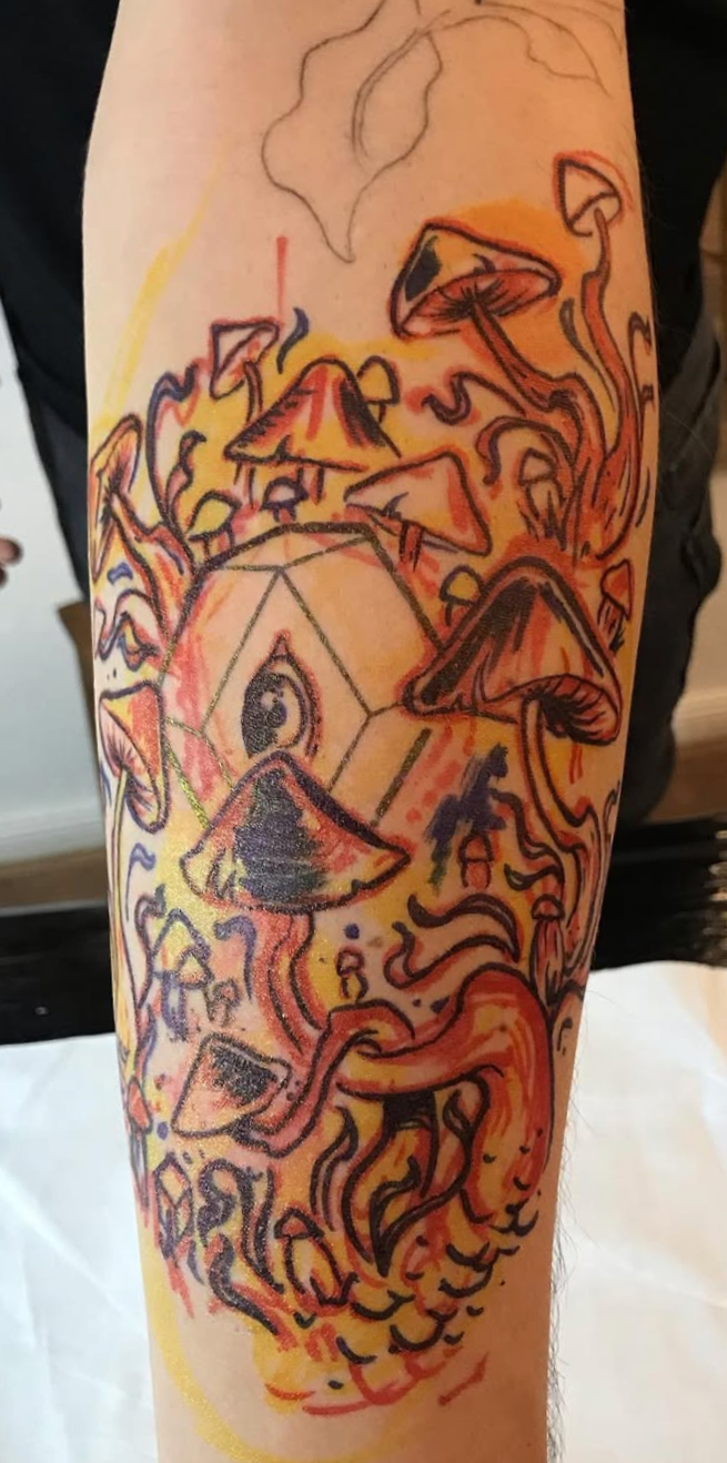 A colorful tattoo on a person's arm featuring mushrooms, an eye within a geometric shape, and abstract swirling lines and shapes.