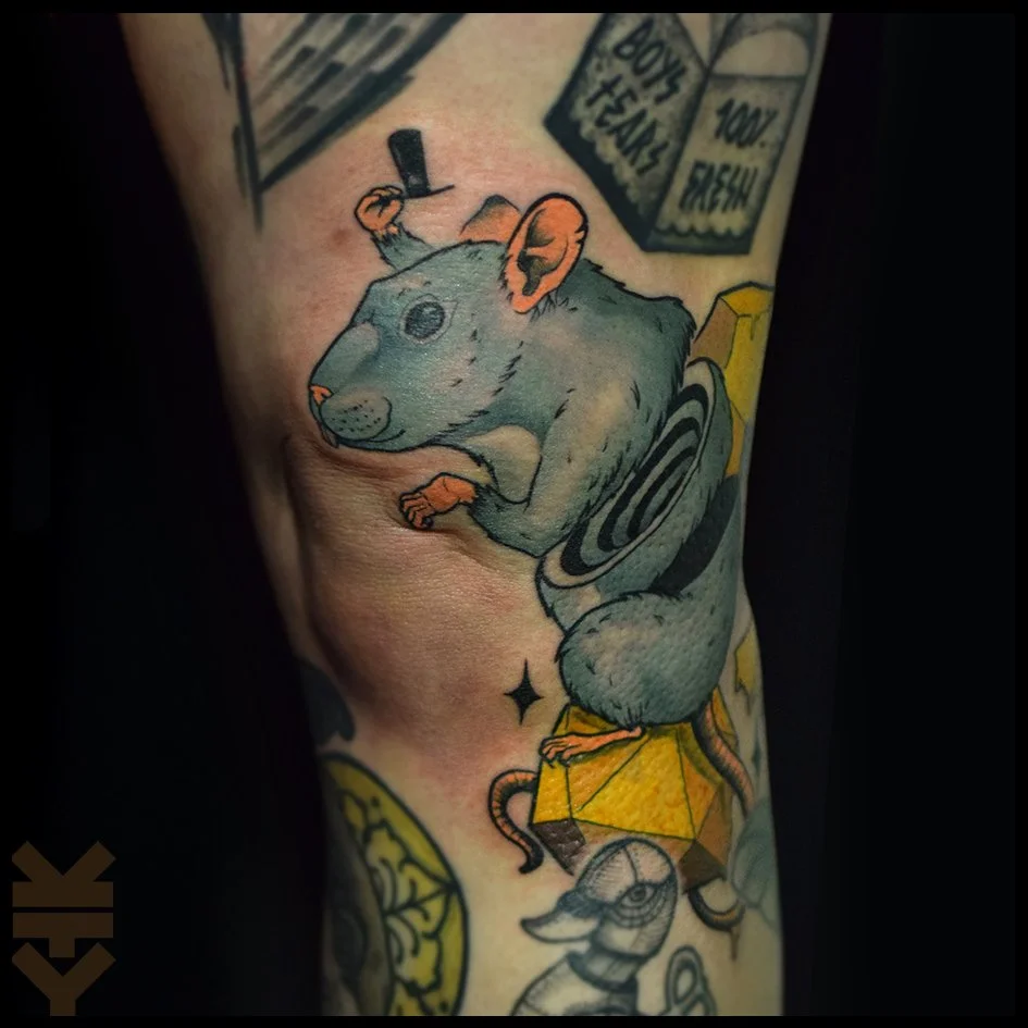 Tattoo of a mouse with a gray body, pink ears, and black eyes, holding a black object on its head, standing on a yellow gemstone, with other elements like dice and a skeleton near it.