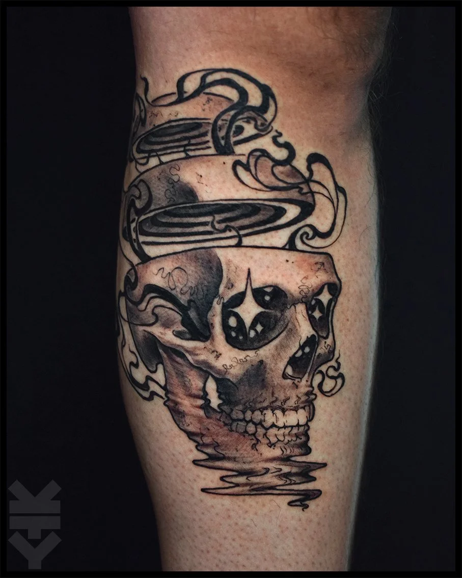 Tattoo of a skull with dark eye sockets and a cracked forehead, with smoke or water swirling above it, in black and gray ink.