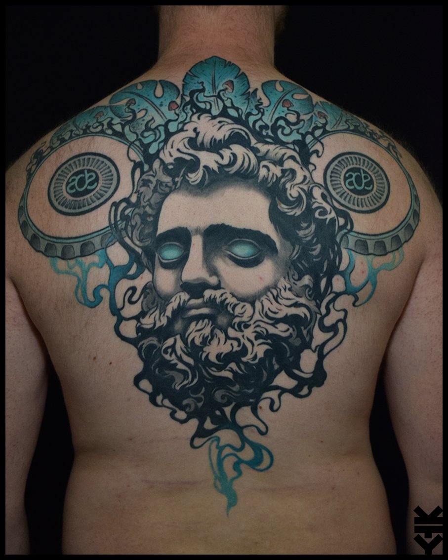 Tattoo of a bearded man's face with glowing eyes, surrounded by smoke, on a person's chest, with speakers and decorative elements in the background.