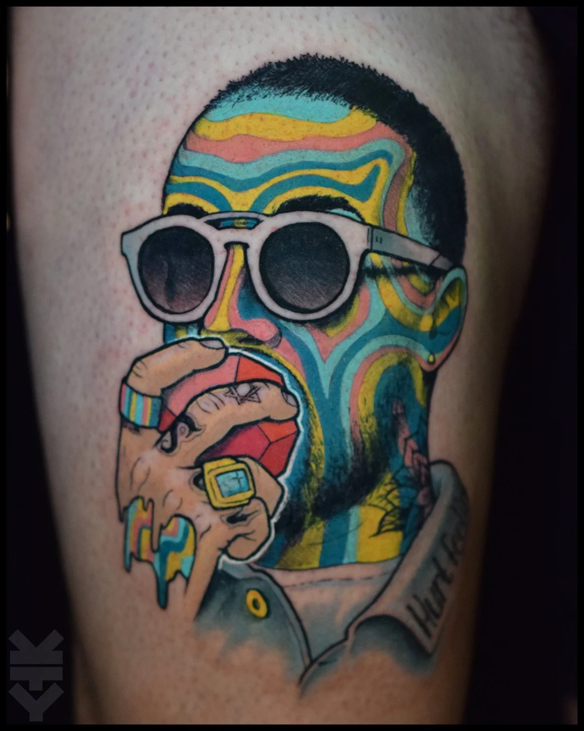 Colorful tattoo of a man wearing sunglasses, holding a red grenade, with rainbow-colored hair and face patterns, and a tattooed shirt. mac miller