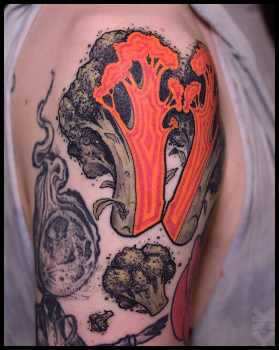 Tattoo of a comic-style broccoli and cauliflower, with bold orange and black outlines on the upper arm.