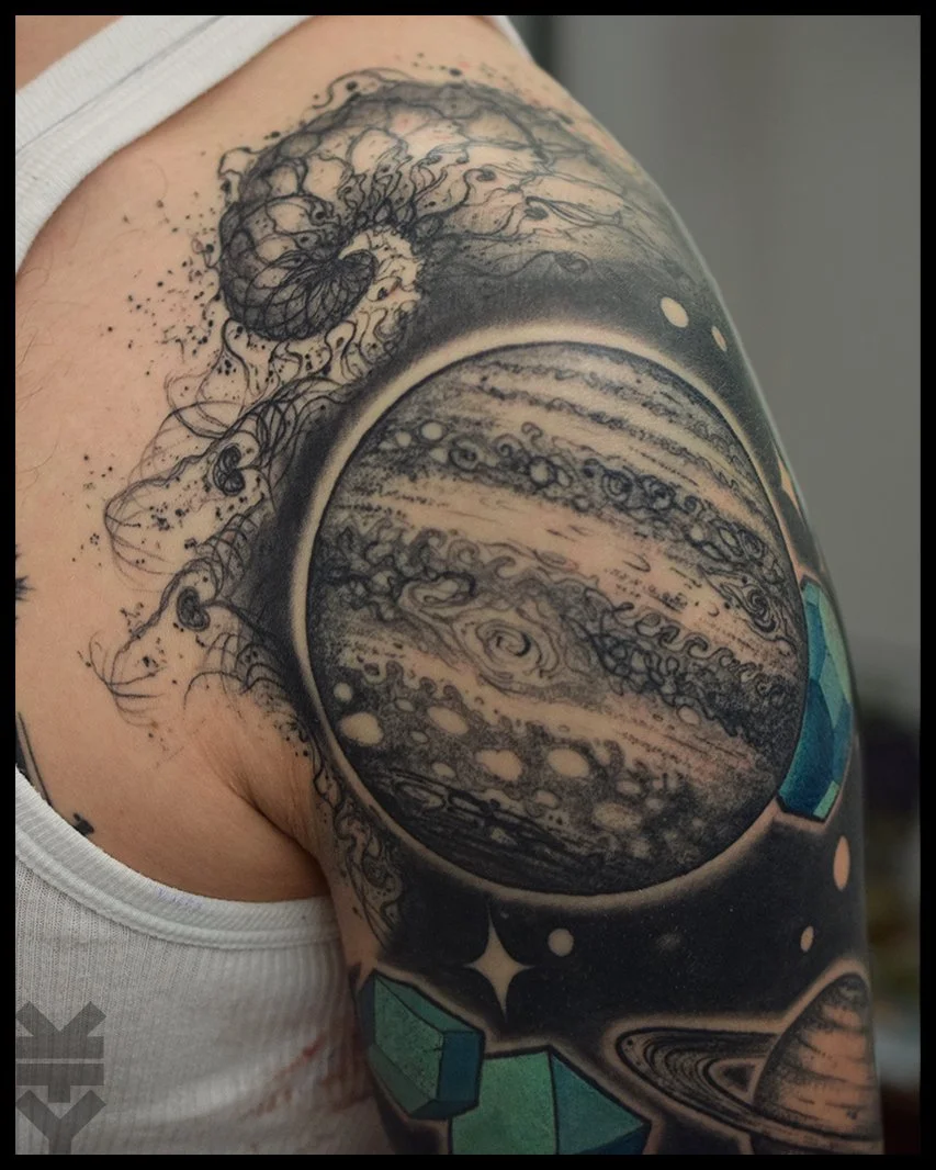 Tattoo of the planet Jupiter with its swirling clouds, surrounded by stars, geometric shapes, and deep space elements, on a person's upper arm.