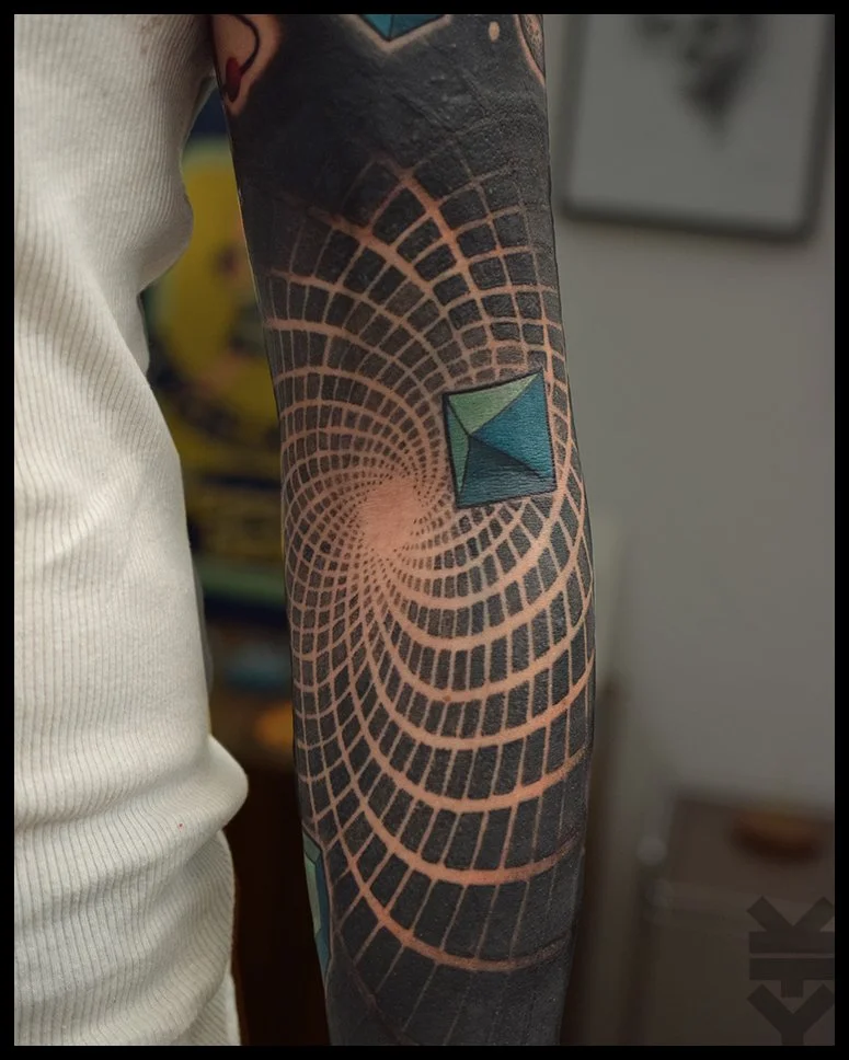 Tattoo of a geometric 3D cube and a spiral pattern on a person's arm, with a fabric sleeve visible on the left.