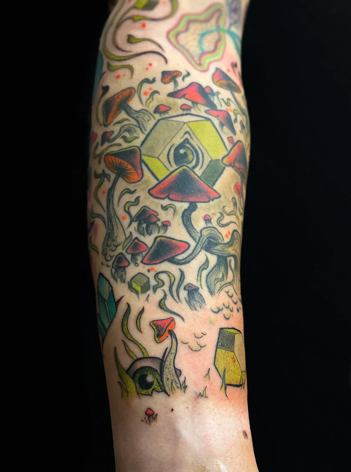 A tattoo on a forearm featuring a colorful, surreal design with mushrooms, an eye within a geometric shape, and various abstract elements.