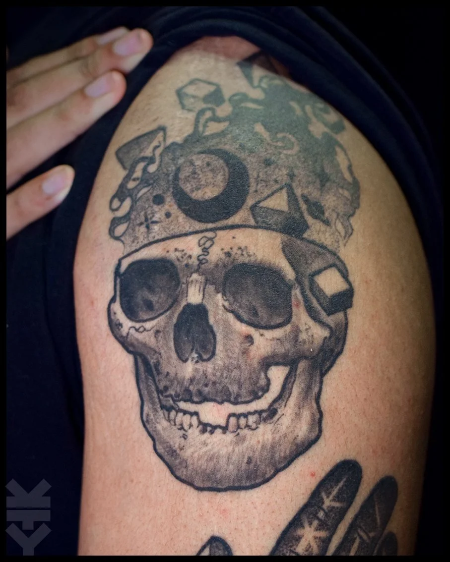 Tattoo of a smiling skull with a top hat featuring a crescent moon, geometric shapes, and abstract designs on skin.