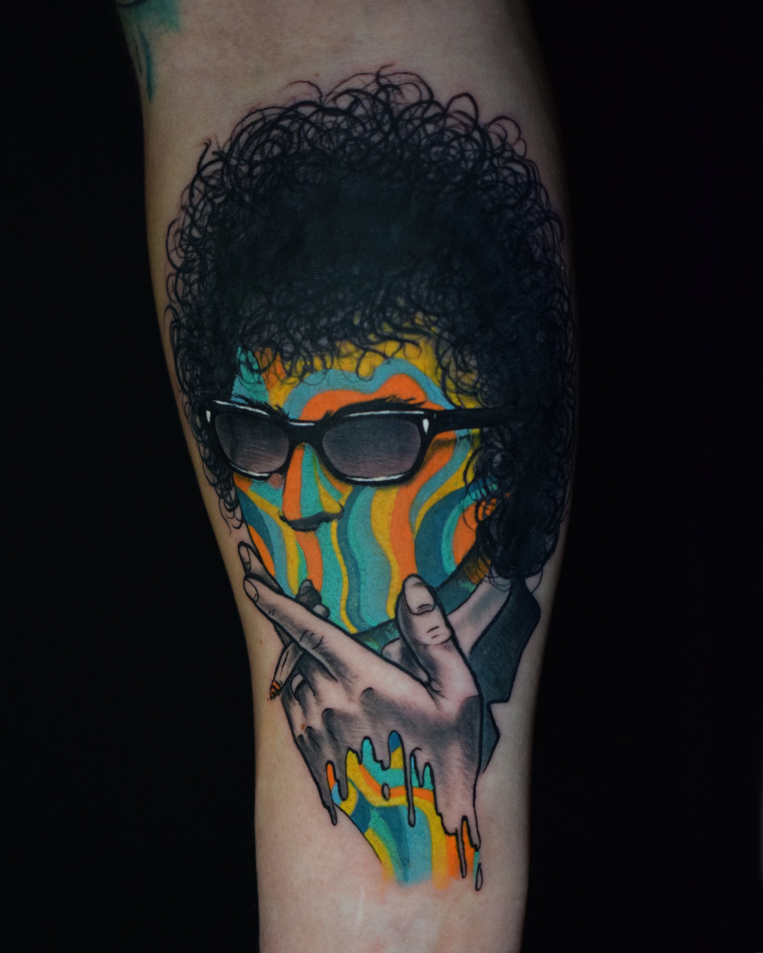 Tattoo of a person with curly hair wearing sunglasses, with colorful abstract behind and a hand holding a joint.