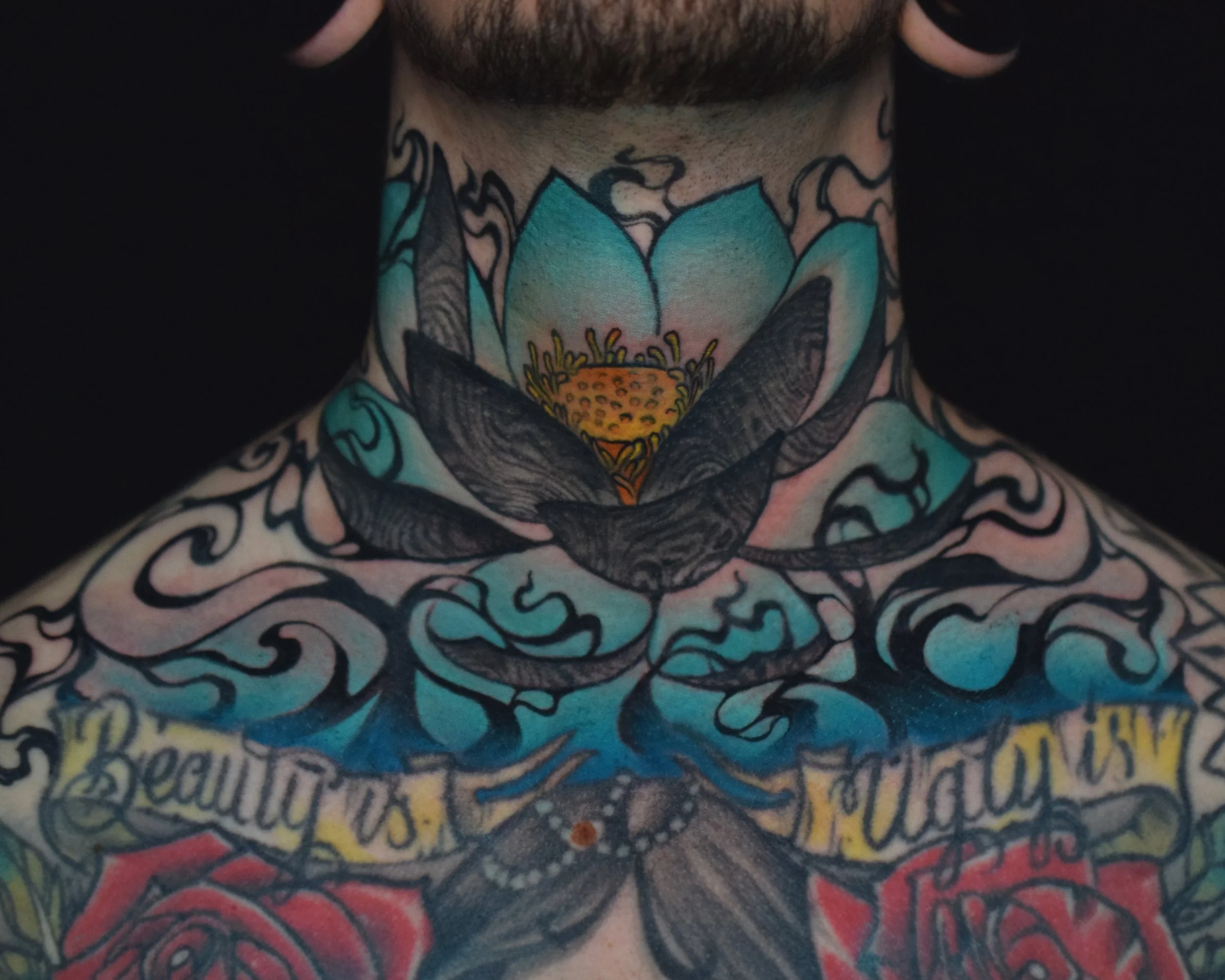 Close-up of a person's chest and neck with colorful tattoos, including a large lotus flower, swirling designs, and the words 'Beauty' and 'Hope' in ornate script.