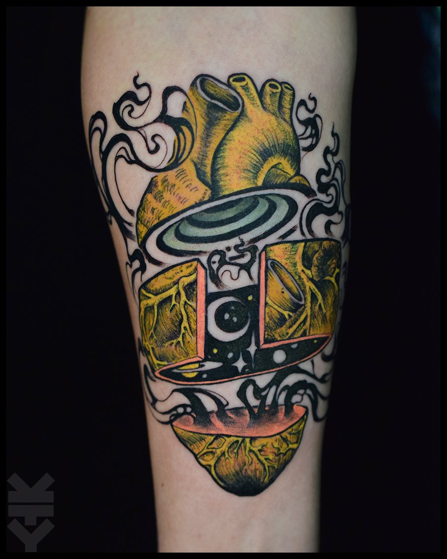 Tattoo of a human heart above a stylized, geometric face with cosmic elements, surrounded by smoke or steam effects.