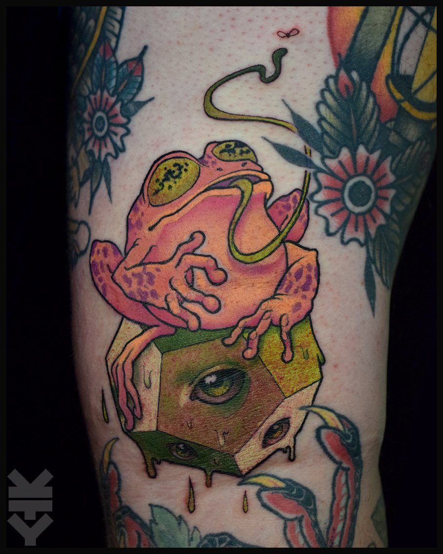 A tattoo of a colorful frog sitting on a cube with eyes painted on it, surrounded by flowers and leaves.