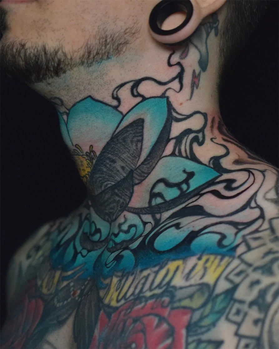 Close-up of a person's neck and chest with colorful tattoos, including a large blue and black flower and swirling designs.