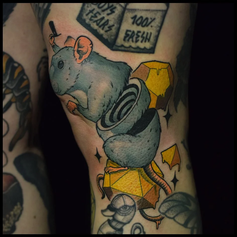 Tattoo of a gray mouse with pink ears and black eyes, sitting on a yellow and brown geometric shape, surrounded by falling yellow pieces, with other tattoos visible on the arm.
