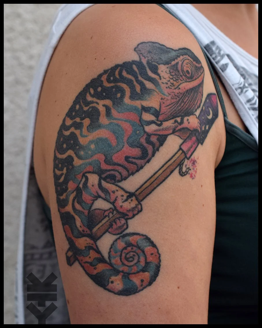 Colorful tattoo of a chameleon holding a paintbrush on upper arm.
