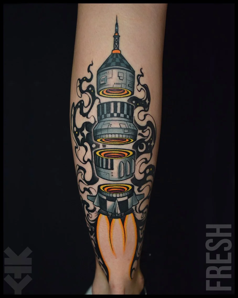 A tattoo of a rocket ship launching on a person's forearm, with black and gray smoke and flames coming from the rocket's engines.