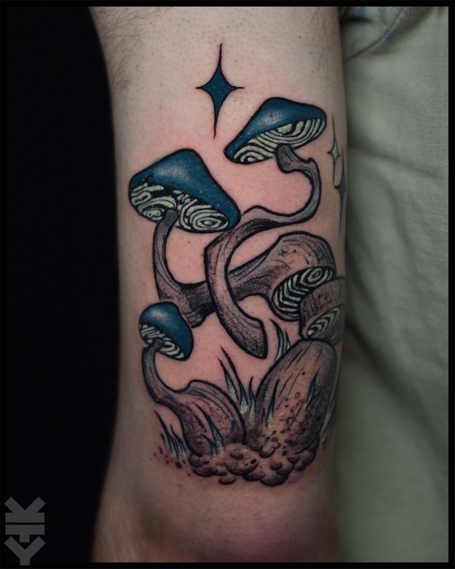 Tattoo of blue and black mushrooms with stylized caps and stems growing from a patch of ground.