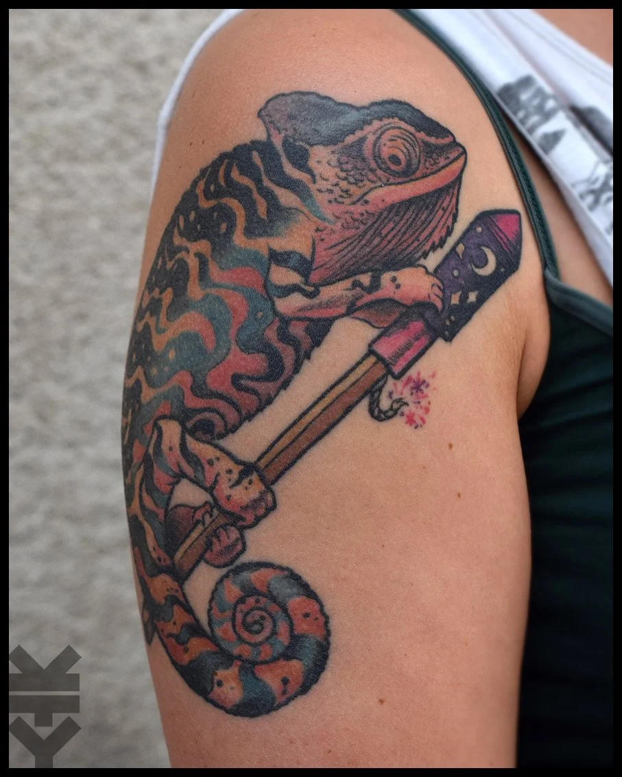 Tattoo of a chameleon holding a hammer with a pink handle, designed with black, orange, and red colors, on a person's upper arm.