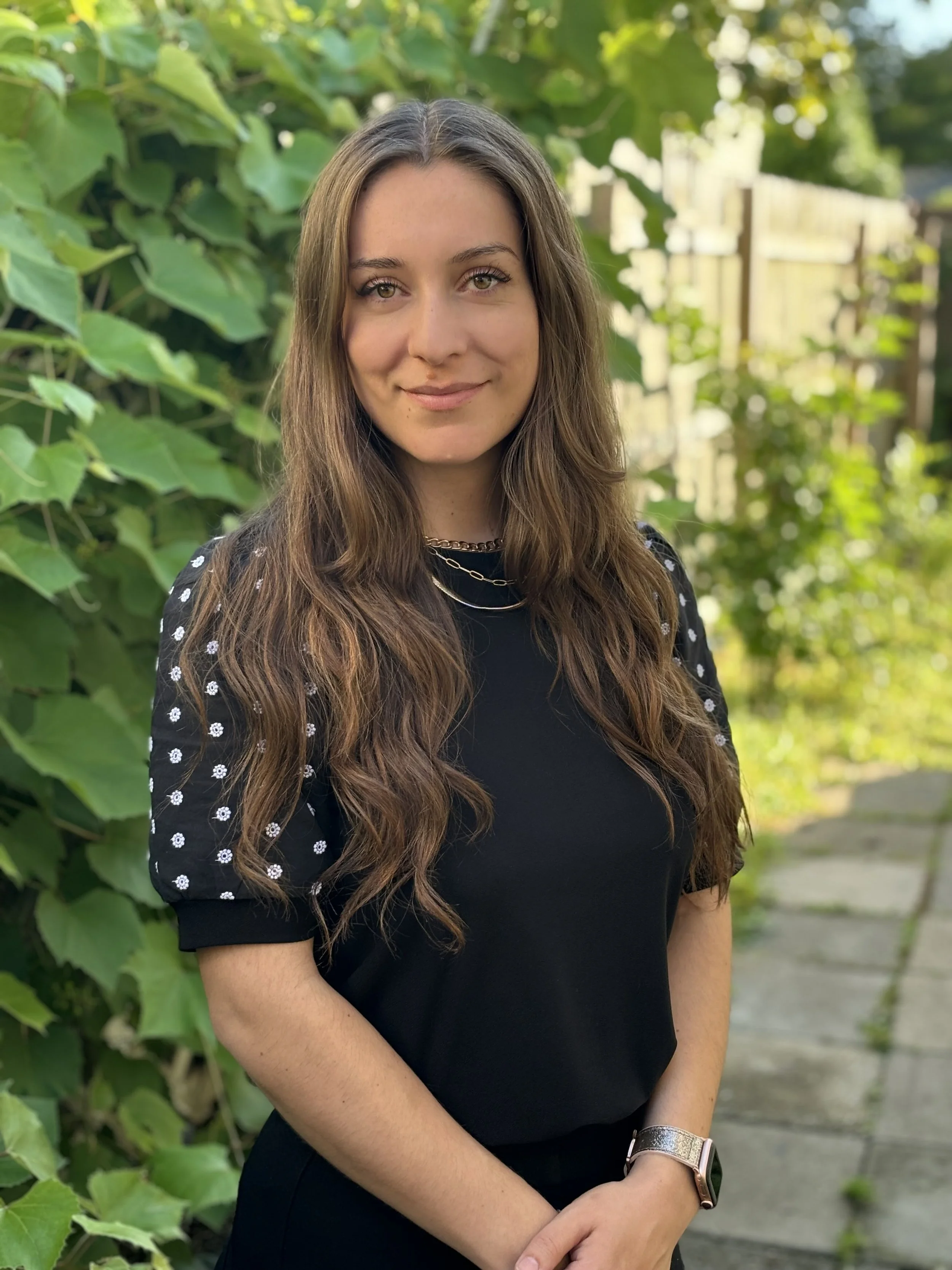 A woman with long wavy brown hair wearing a black top with sheer sleeves decorated with white floral patterns, standing outdoors with green foliage and a wooden fence in the background.