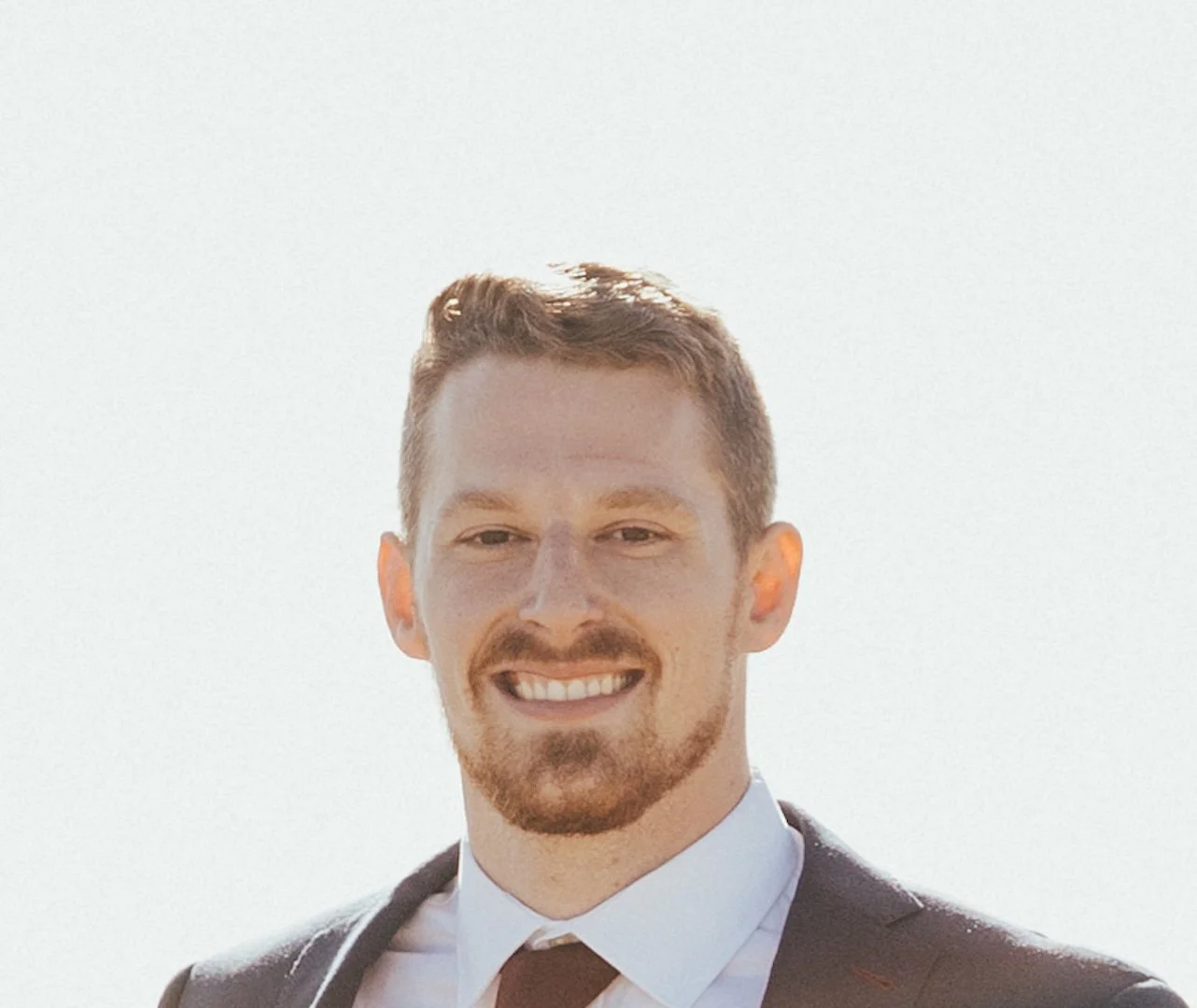 A young man with red hair, a beard, and a mustache smiling outdoors in front of a bright, clear sky. He is wearing a suit and tie.