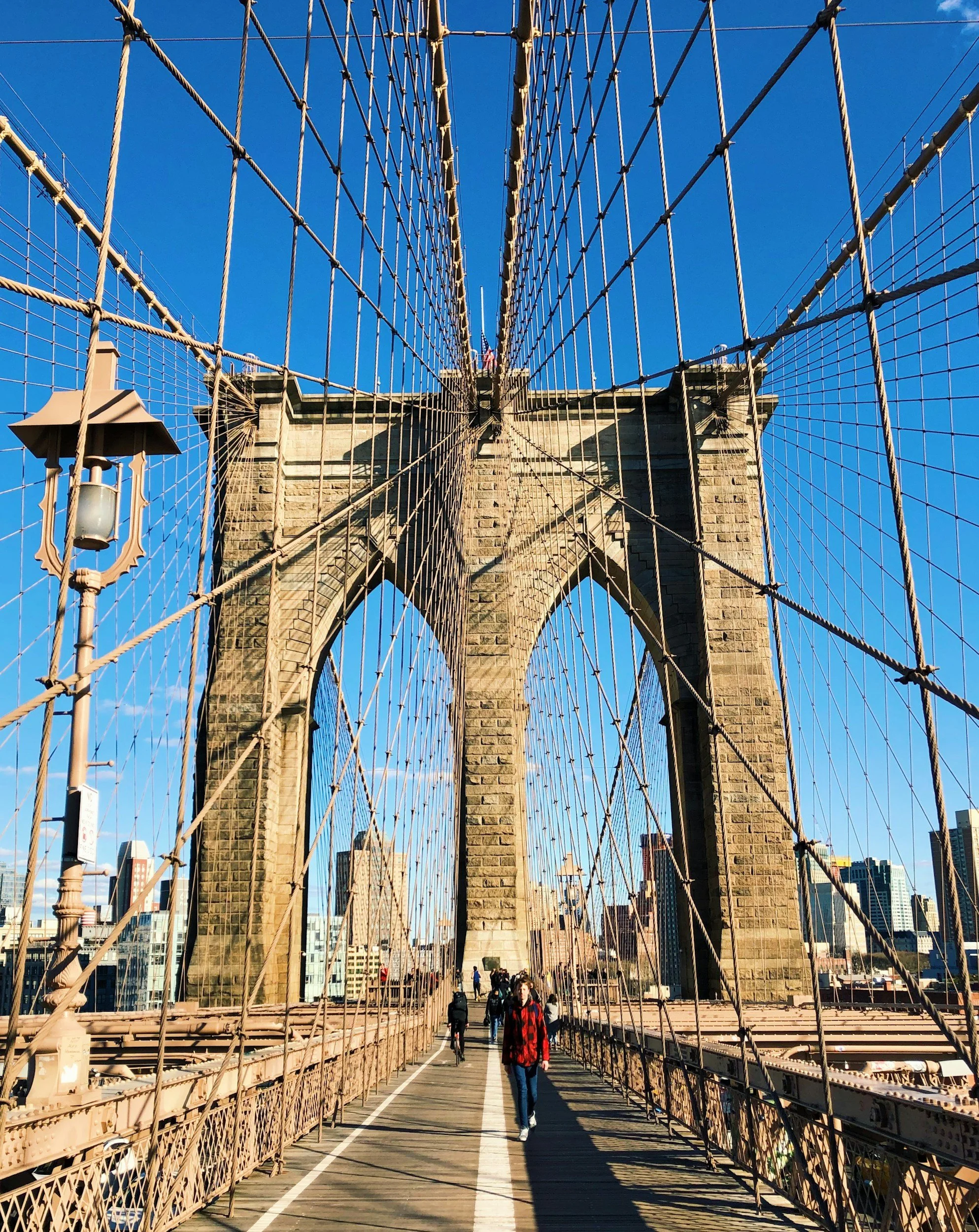 View of Brooklyn Bridge with pedestrians walking on the wooden walkway, perspective looking up at the stone towers and wire suspension cables, New York City skyline in the background, clear blue sky.