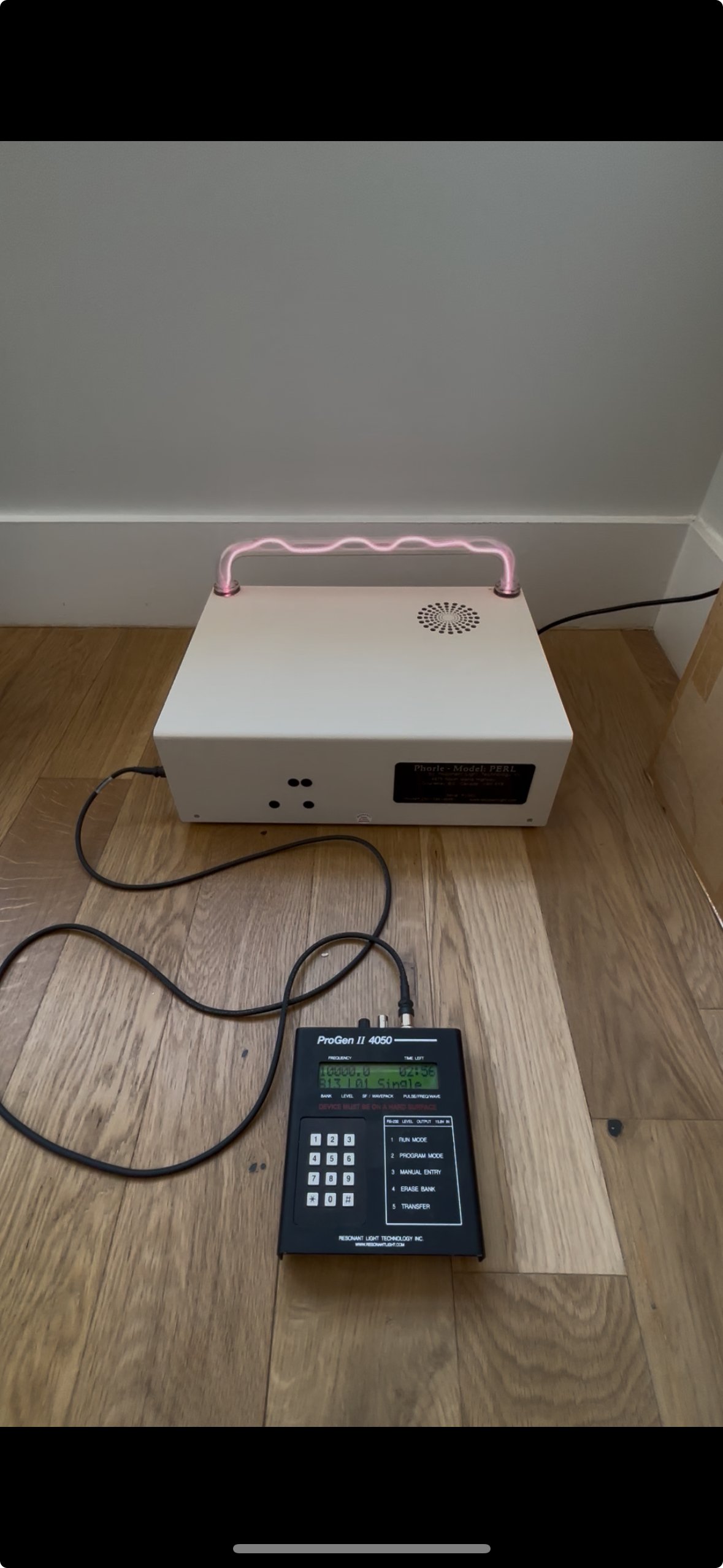 A phototherapy device with a glowing pink neon light handle connected to a control unit with a digital display, placed on a wooden floor against a wall.