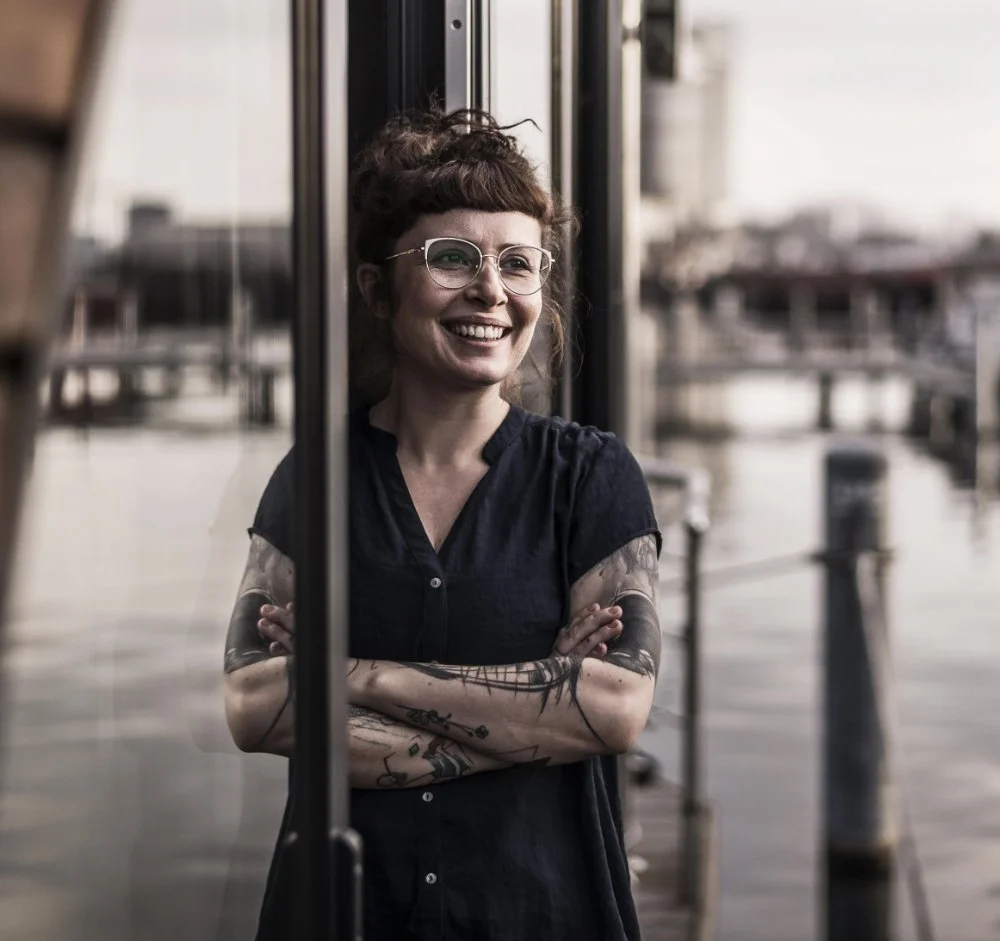 Eine lachende Frau mit Tattoos, trägt eine schwarze Bluse und trägt Brillen, steht an einem Fenster an einem Hafen