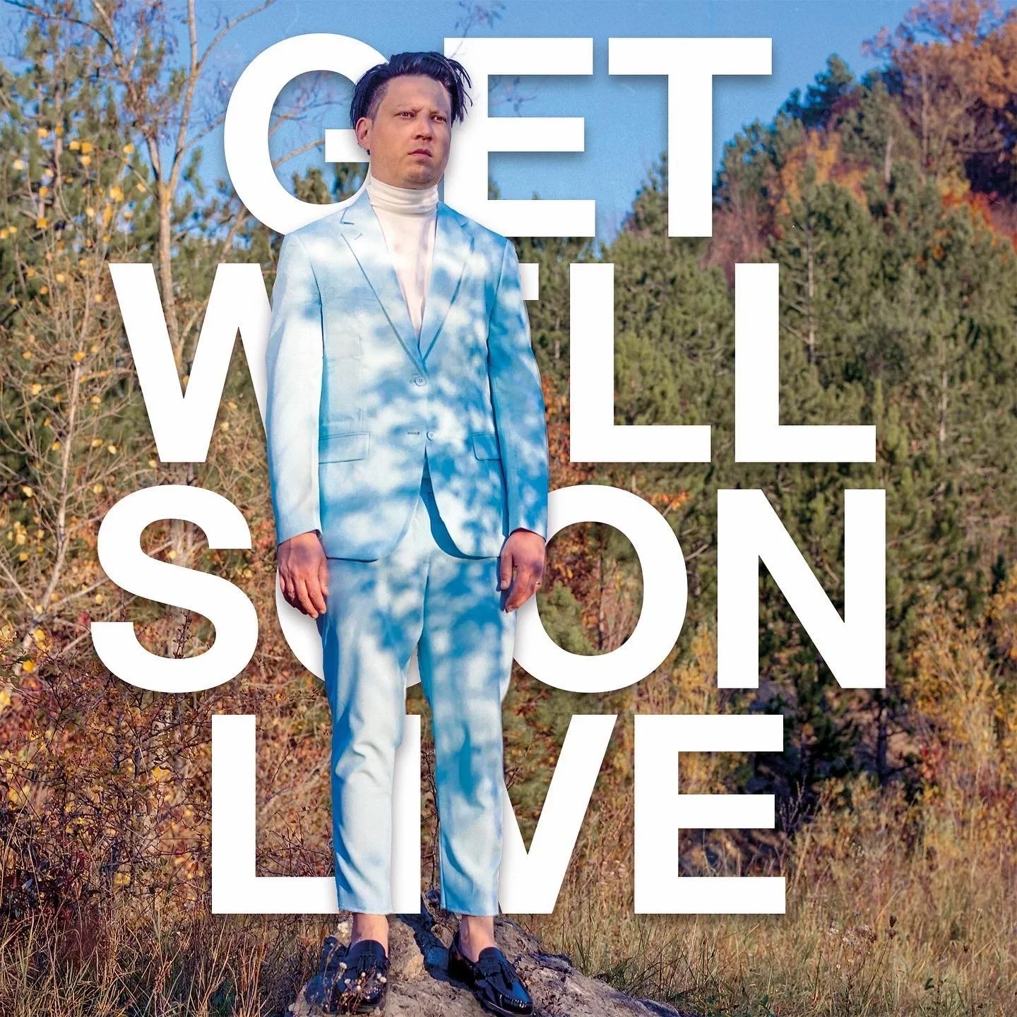 Ein Mann in einem hellblauen Anzug steht im Freien vor Bäumen mit herbstlicher Laubfärbung. Über ihm sind große weiße Buchstaben, die den Titel „Gret Willson Live“ bilden.