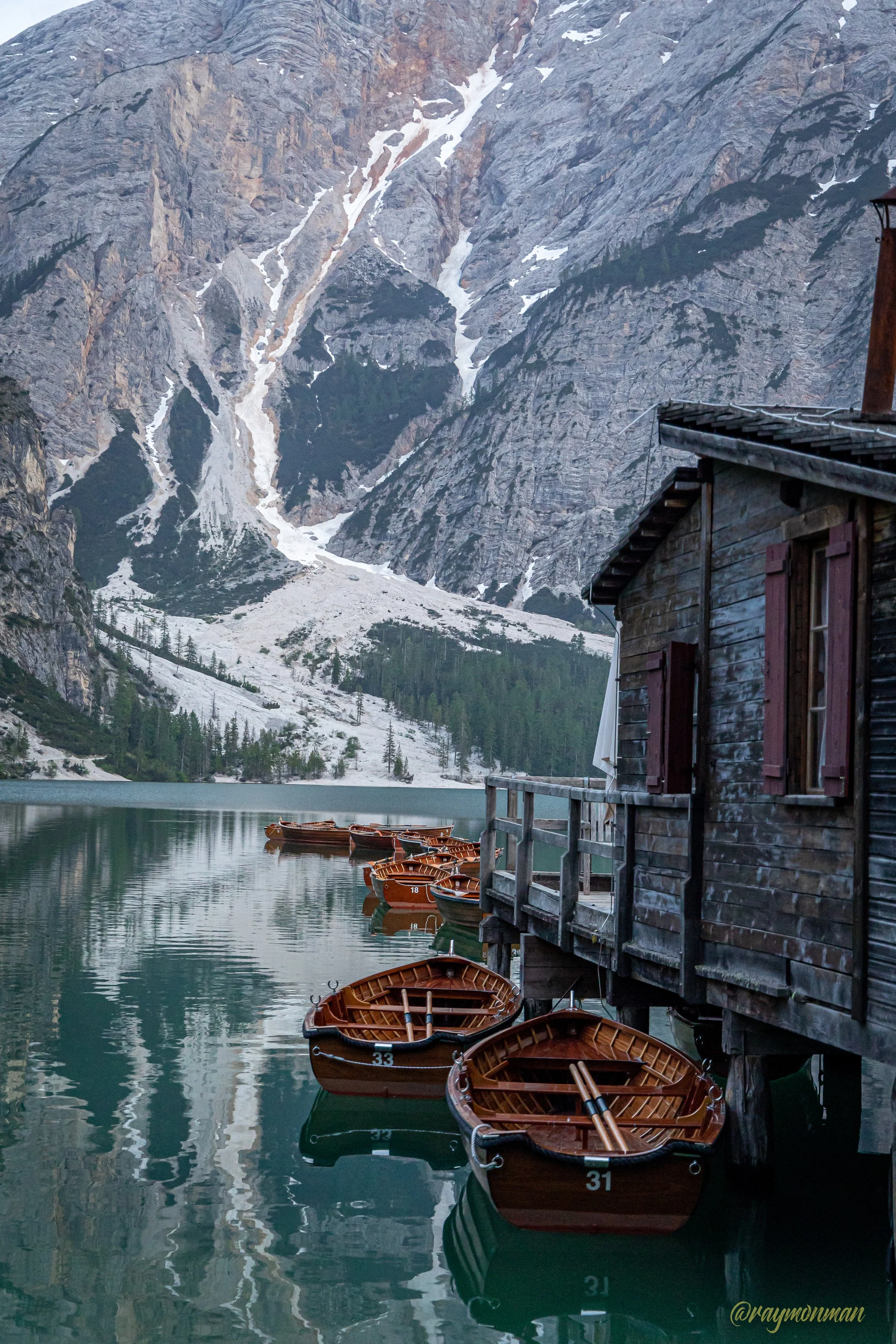 Vista de un lago rodeado de montañas con nieve y árboles verdes, con varias barcas de madera ancladas y un cobertizo de madera a la orilla del agua.