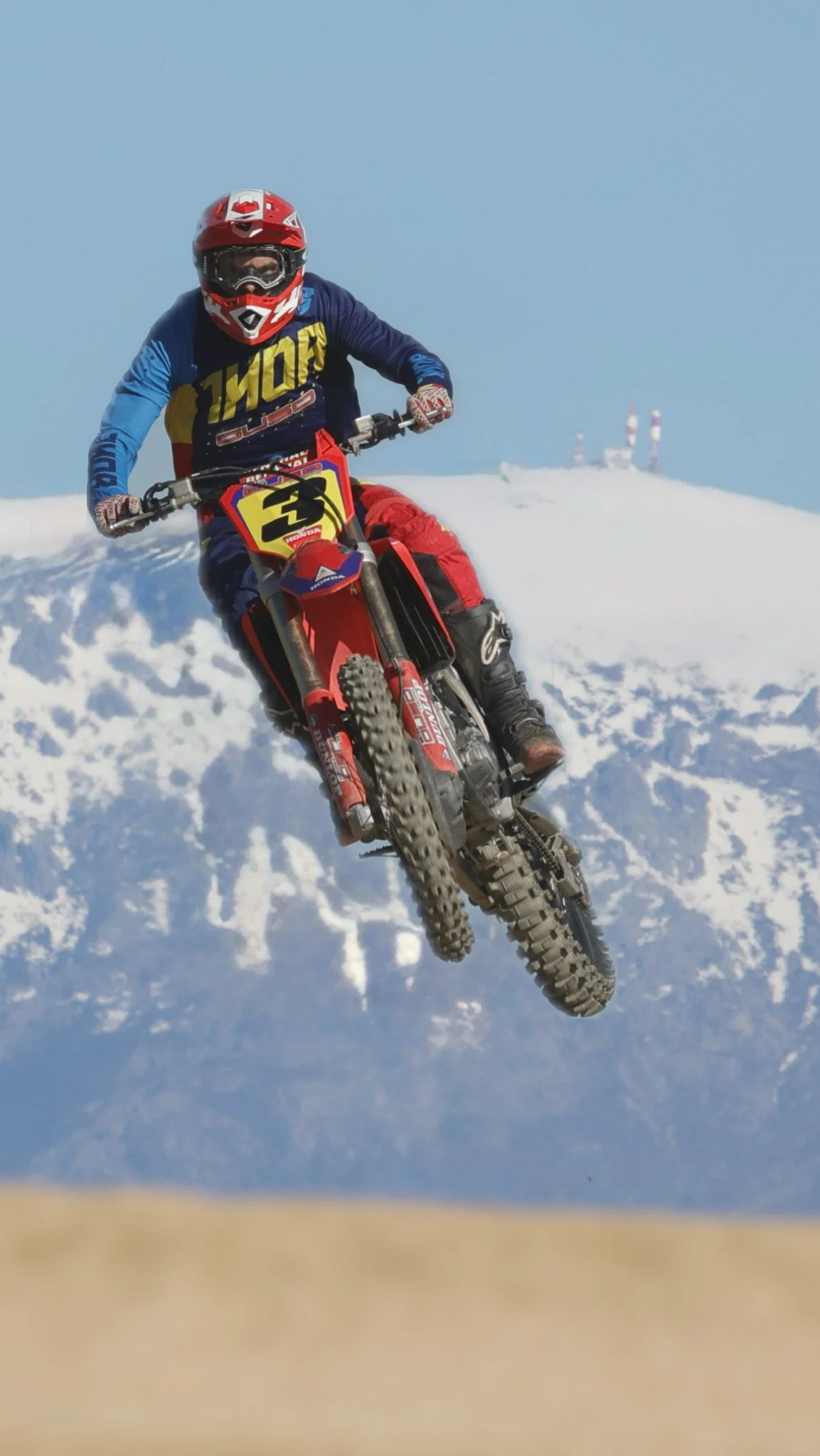 Un ciclista en moto de dirt biking saltando en una montaña nevada con picos y radiobases en el fondo.