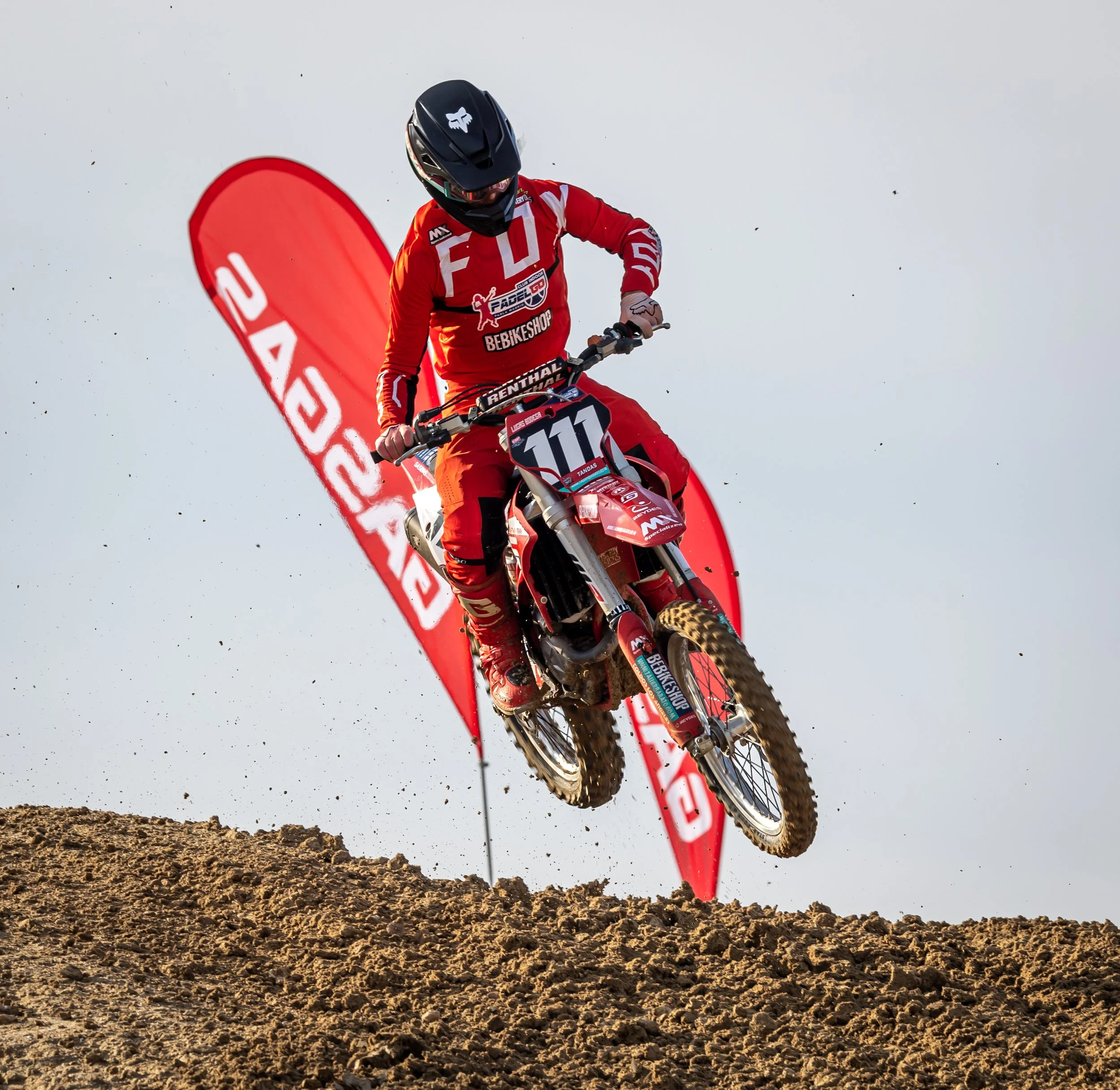 Motociclista en traje rojo, con casco negro, saltando sobre tierra durante una carrera de motocross, con banderas rojas de fondo que dicen 'JACKED'.