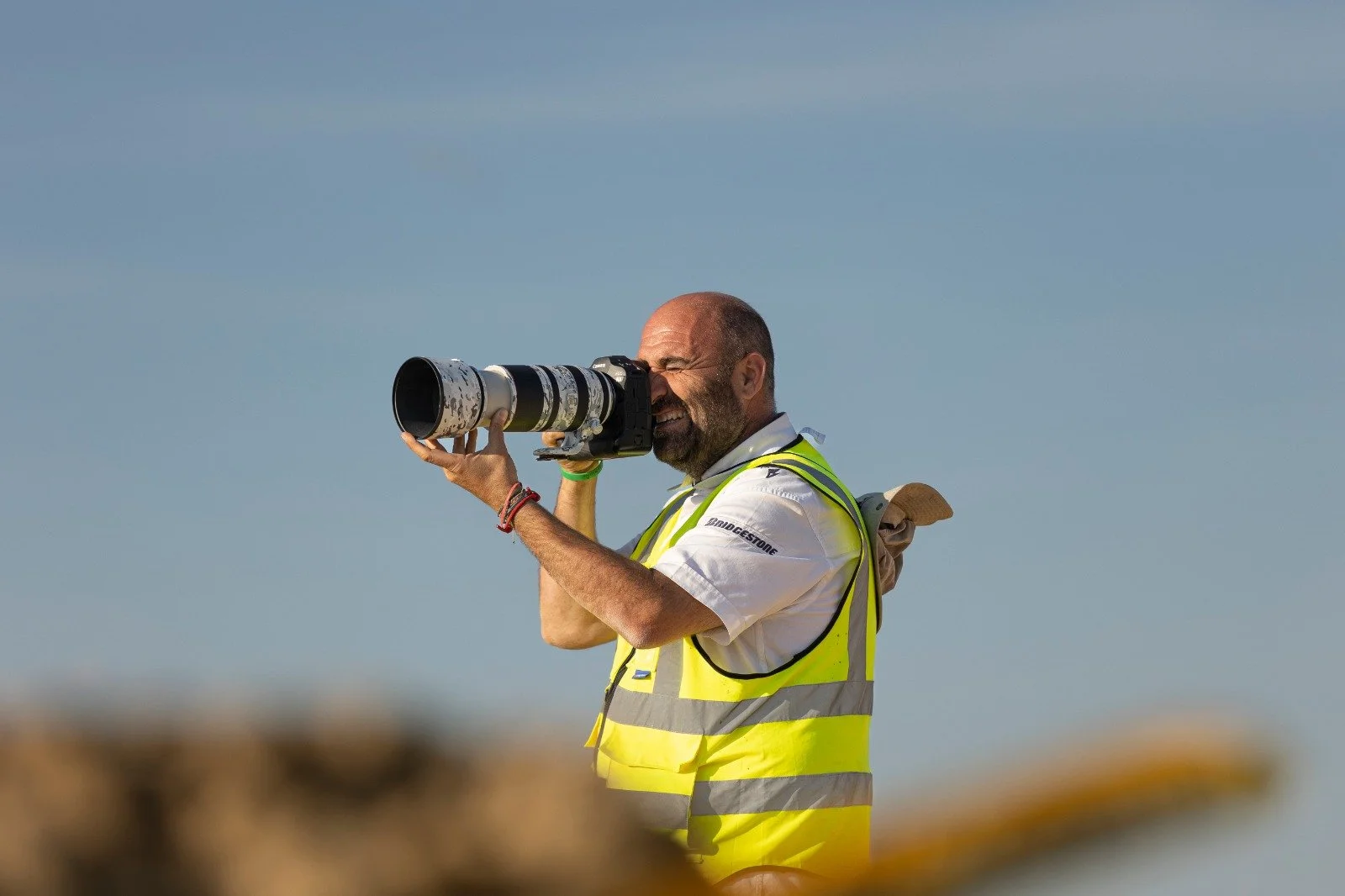 Hombre con chaleco de seguridad, sosteniendo un teleobjetivo, fotografiando en un espacio al aire libre. Raymonman