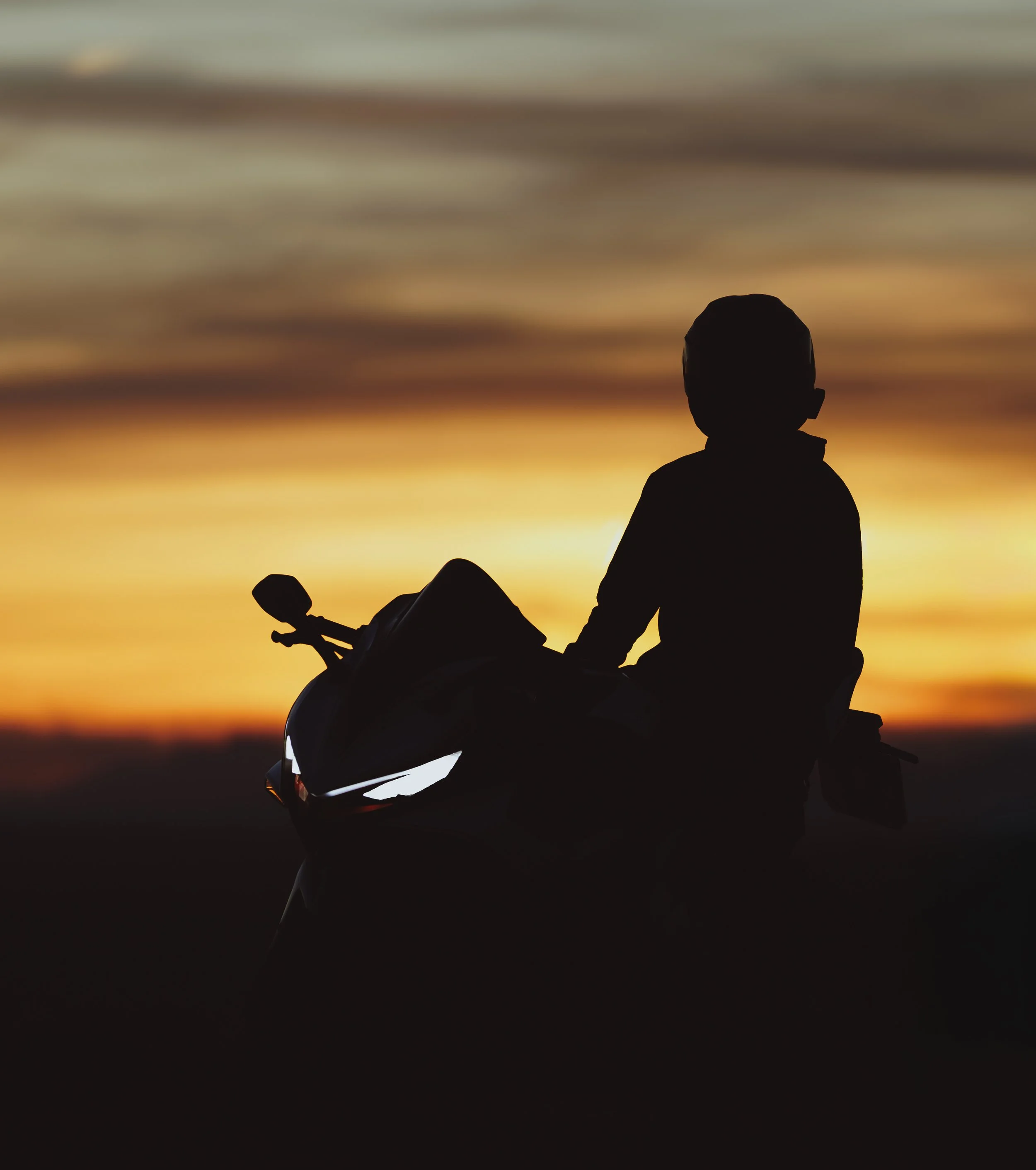Silueta de una persona con casco, sentado en una motocicleta, durante un atardecer con cielo nublado en el fondo.