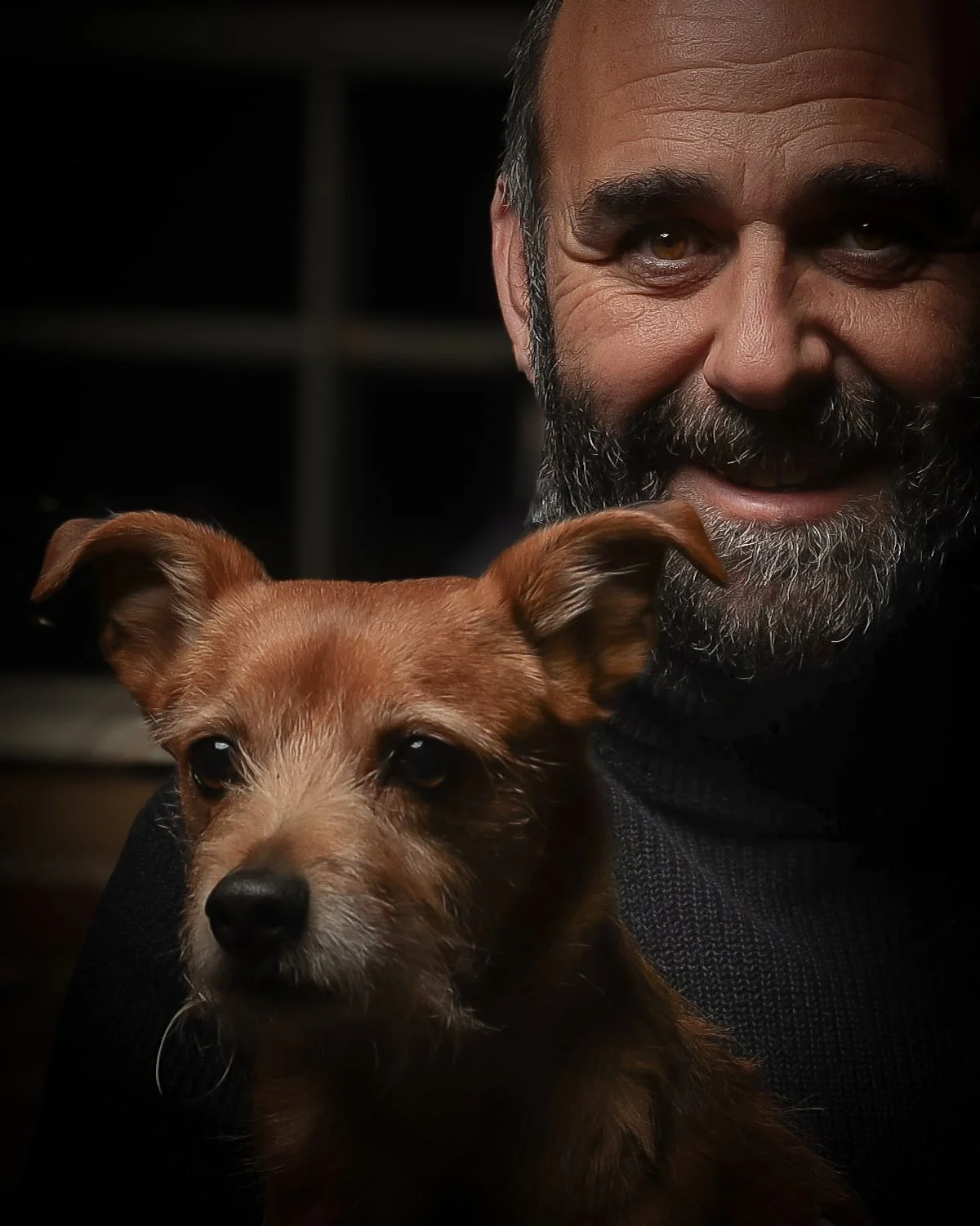 Hombre con barba sonriendo y un perro pequeño de color marrón oscuro en primer plano.