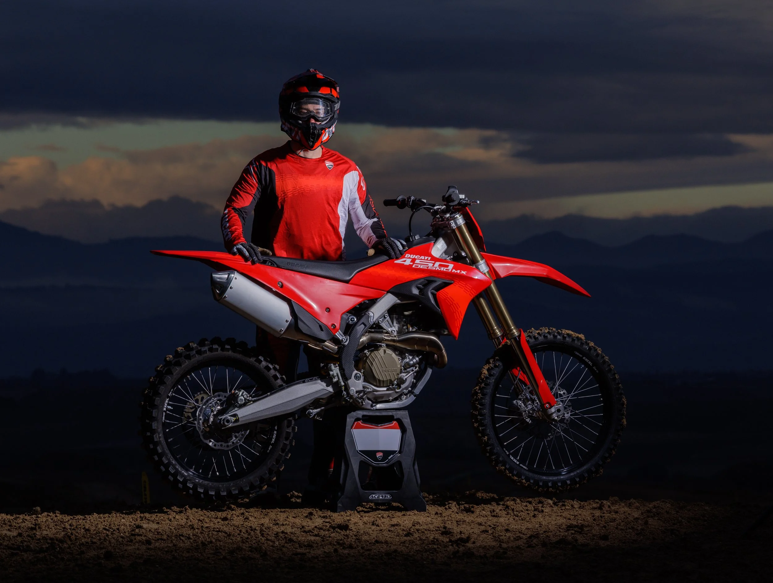 Persona con equipo de protección y casco de motocross, con una motocicleta roja de enduro en un paisaje de montaña al atardecer.