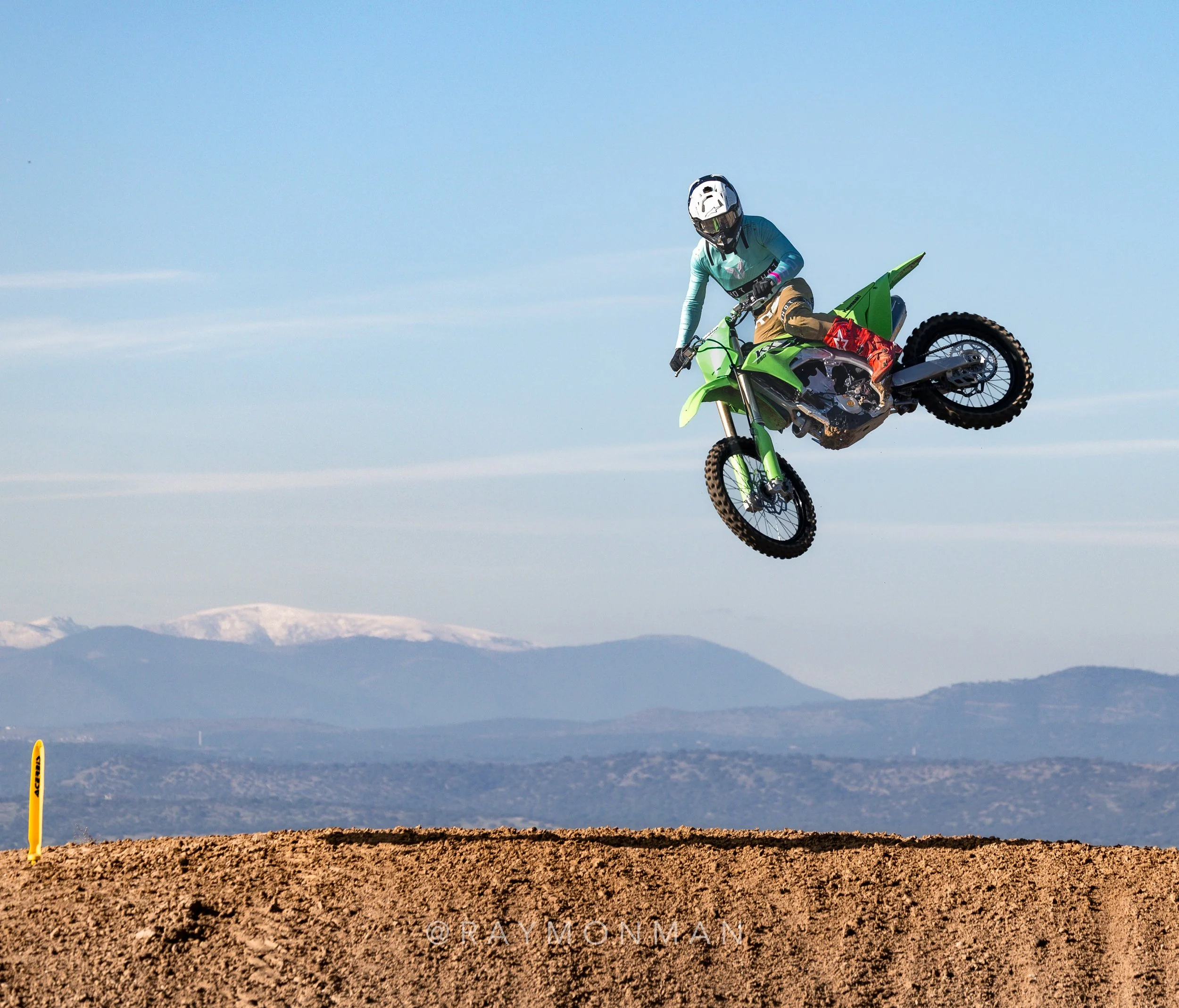 Una persona en motocross saltando en un campo con montañas en el fondo.