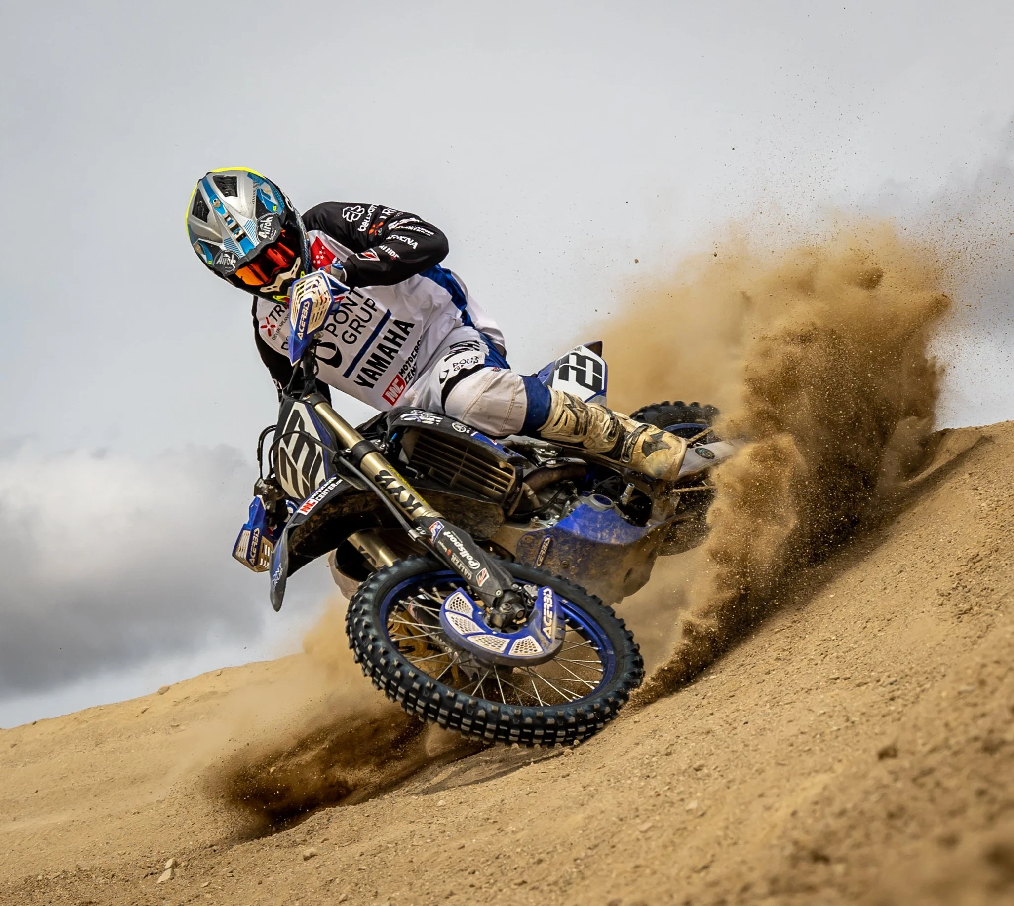 Un piloto de motocross en una moto azul y negra, vestido con equipo de protección y casco, inclinándose mientras conduce a gran velocidad en arena, levantando polvo.