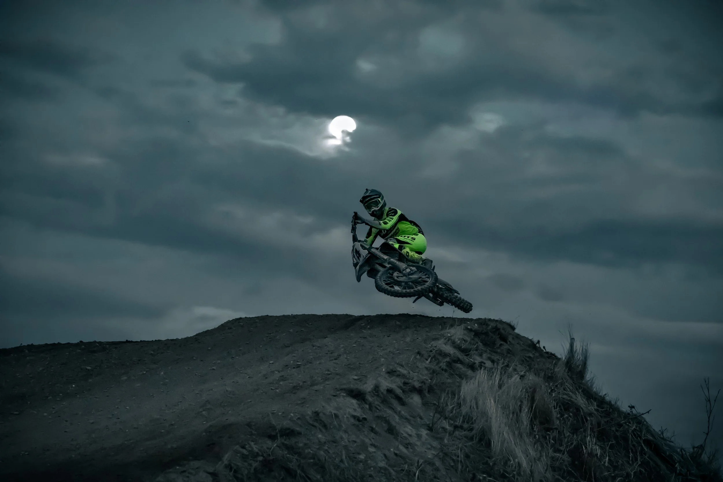 Un motociclista en motocross saltando sobre una pendiente en un paisaje nocturno con cielo nublado y luna parcialmente visible.