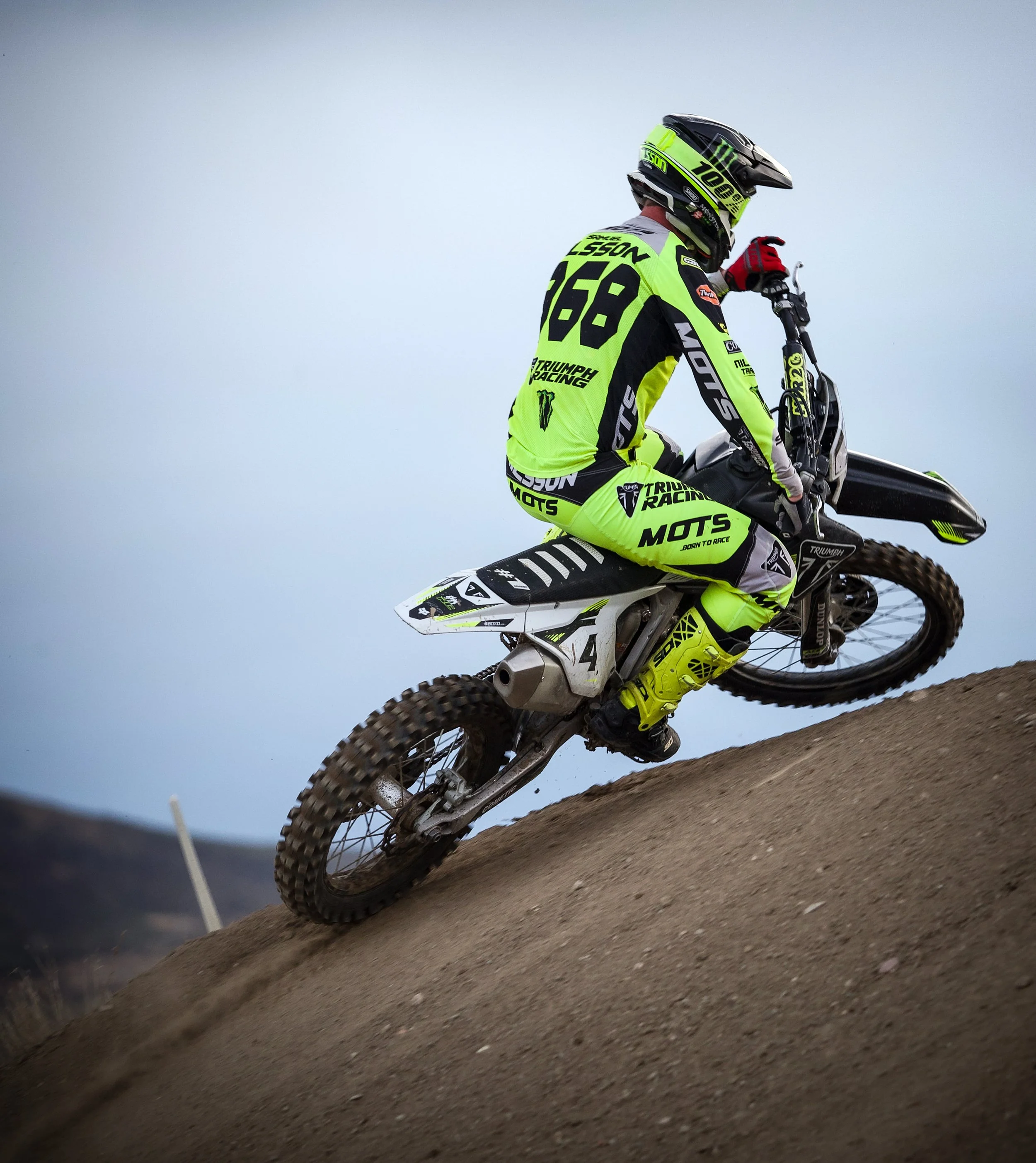 Motociclista vestido con equipo de protección y ropa de color neón, montando una moto de dirt bike en una pendiente inclinada en un terreno arenoso o de tierra, con un cielo despejado al fondo.