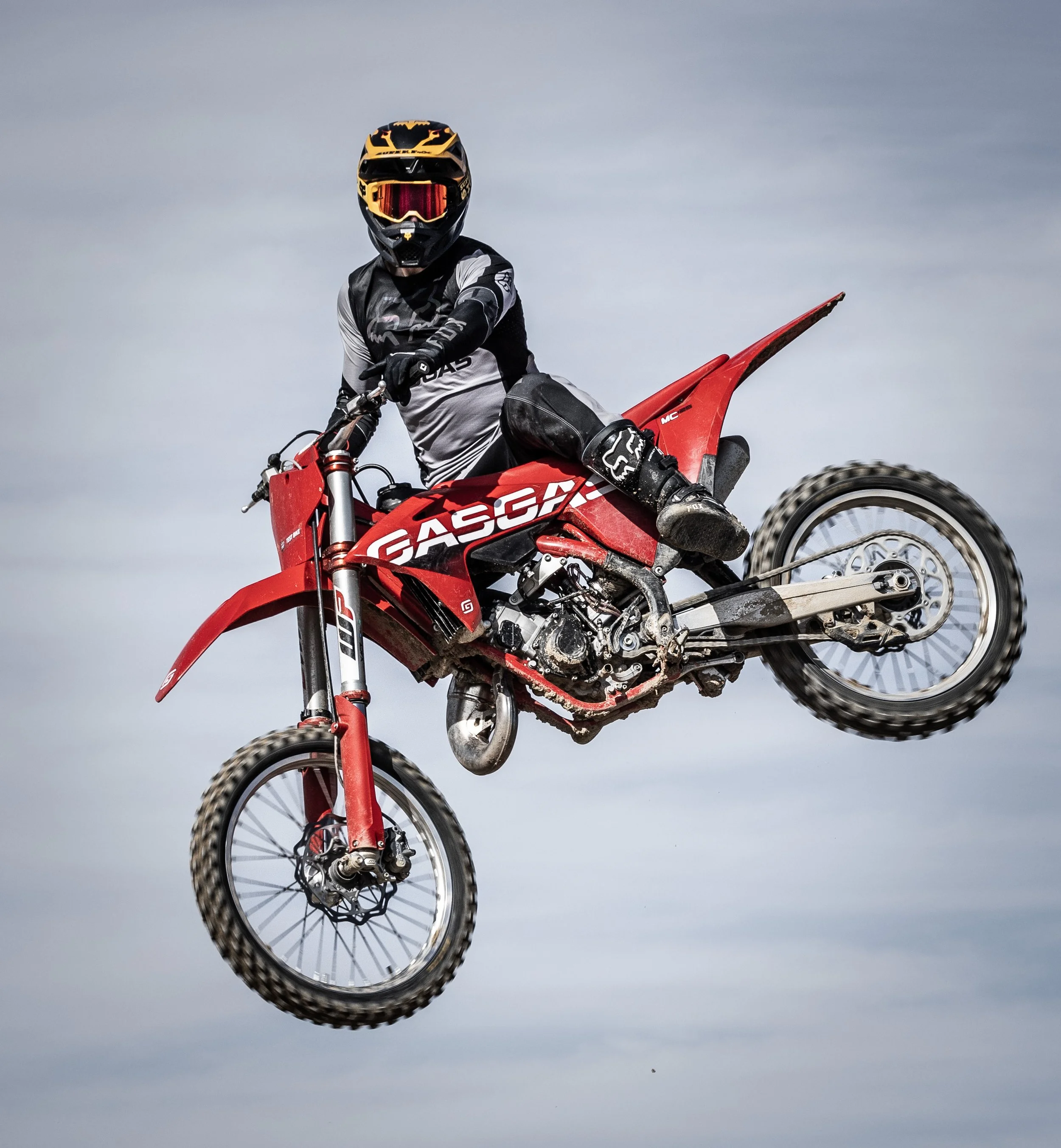 Una persona en un motocicleta de enduro en el aire, con traje de protección, casco y gafas, en un cielo nublado.
