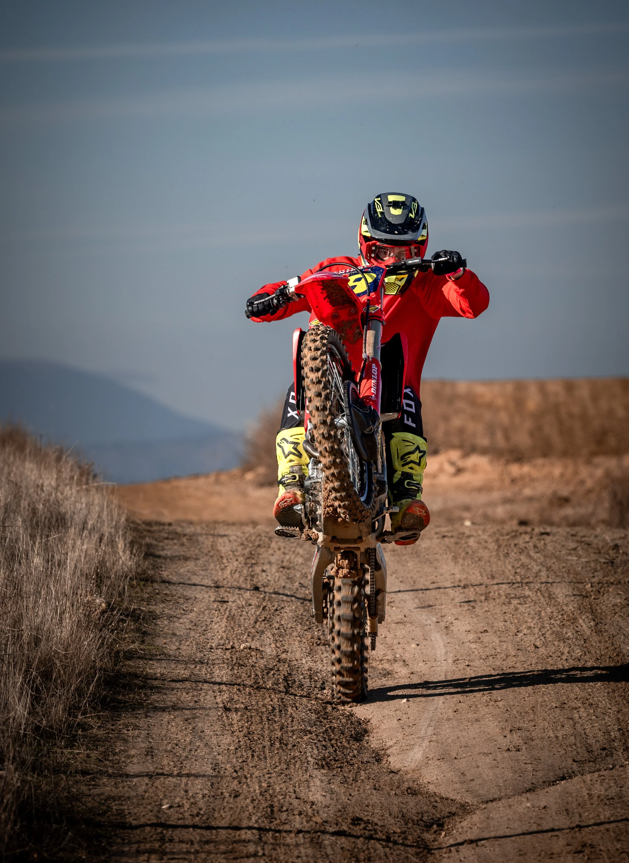Persona haciendo una pirueta en bicicleta de dirt jump en un sendero de tierra en un área abierto con montañas al fondo.
