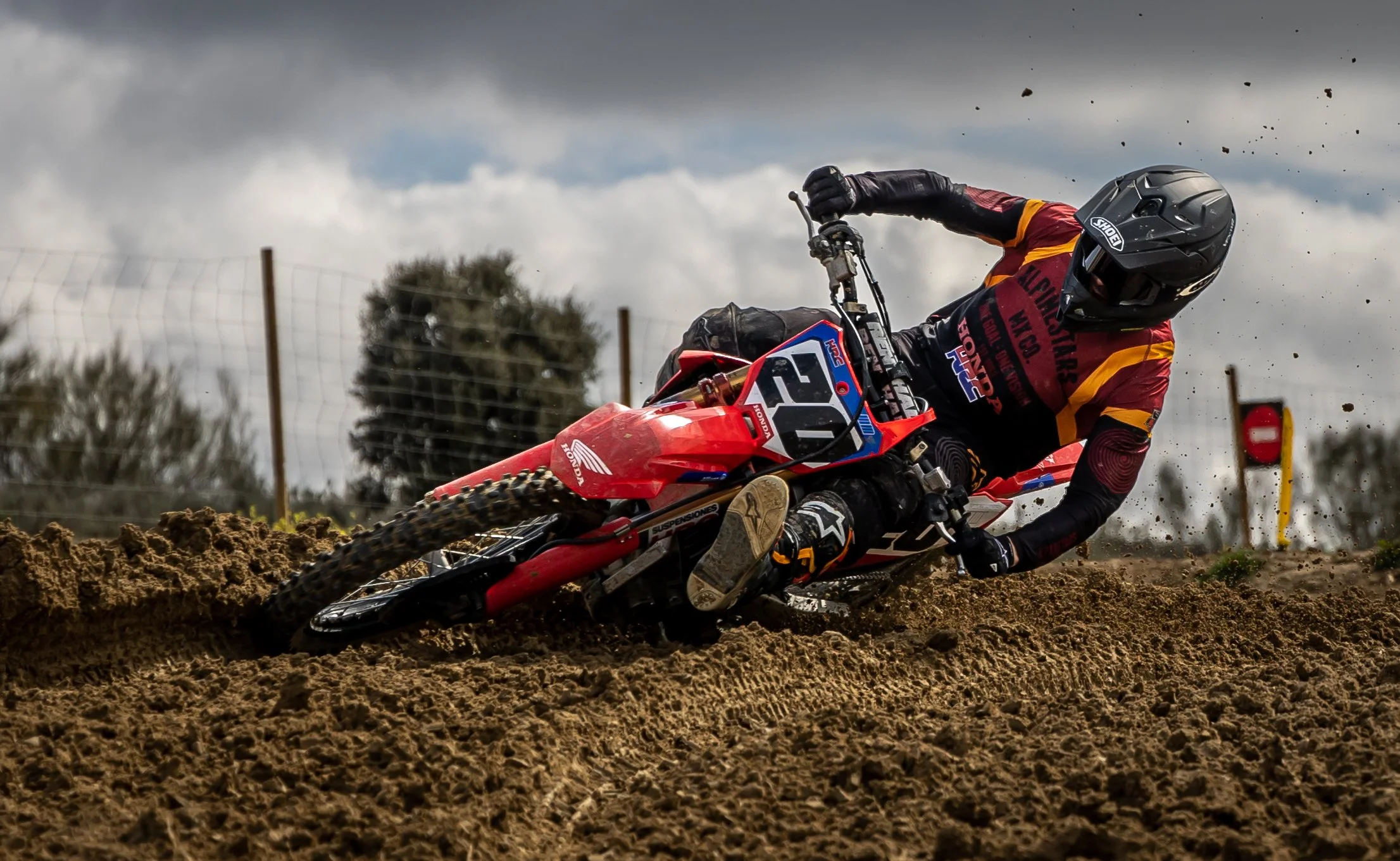 Persona en motocicleta de enduro, usando casco y equipo de protección, maniobrando en un terreno de tierra, con cielo nublado y señal de advertencia en el fondo.