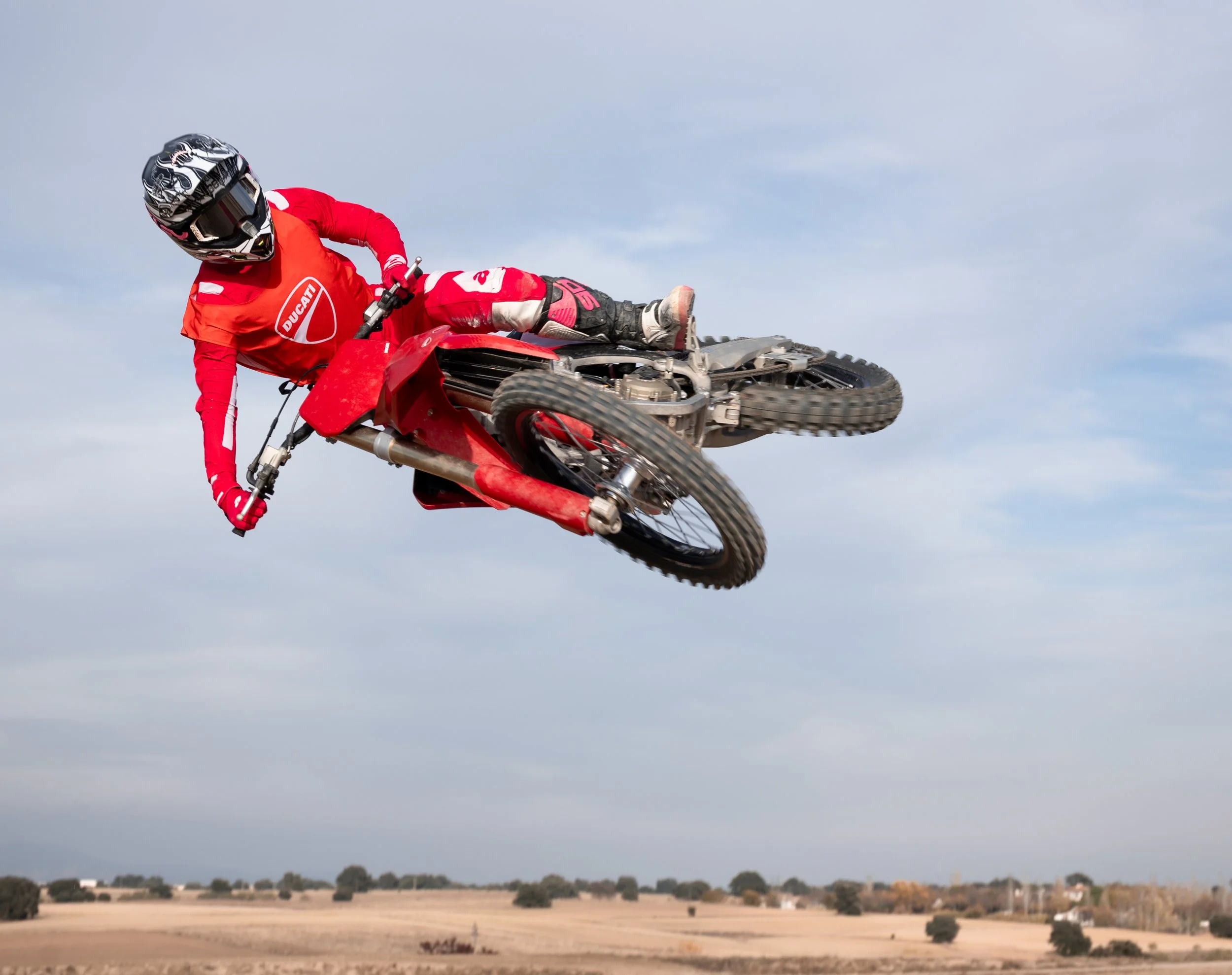 Persona surfeando en una moto de motocross en el aire con campo y cielo nublado al fondo.