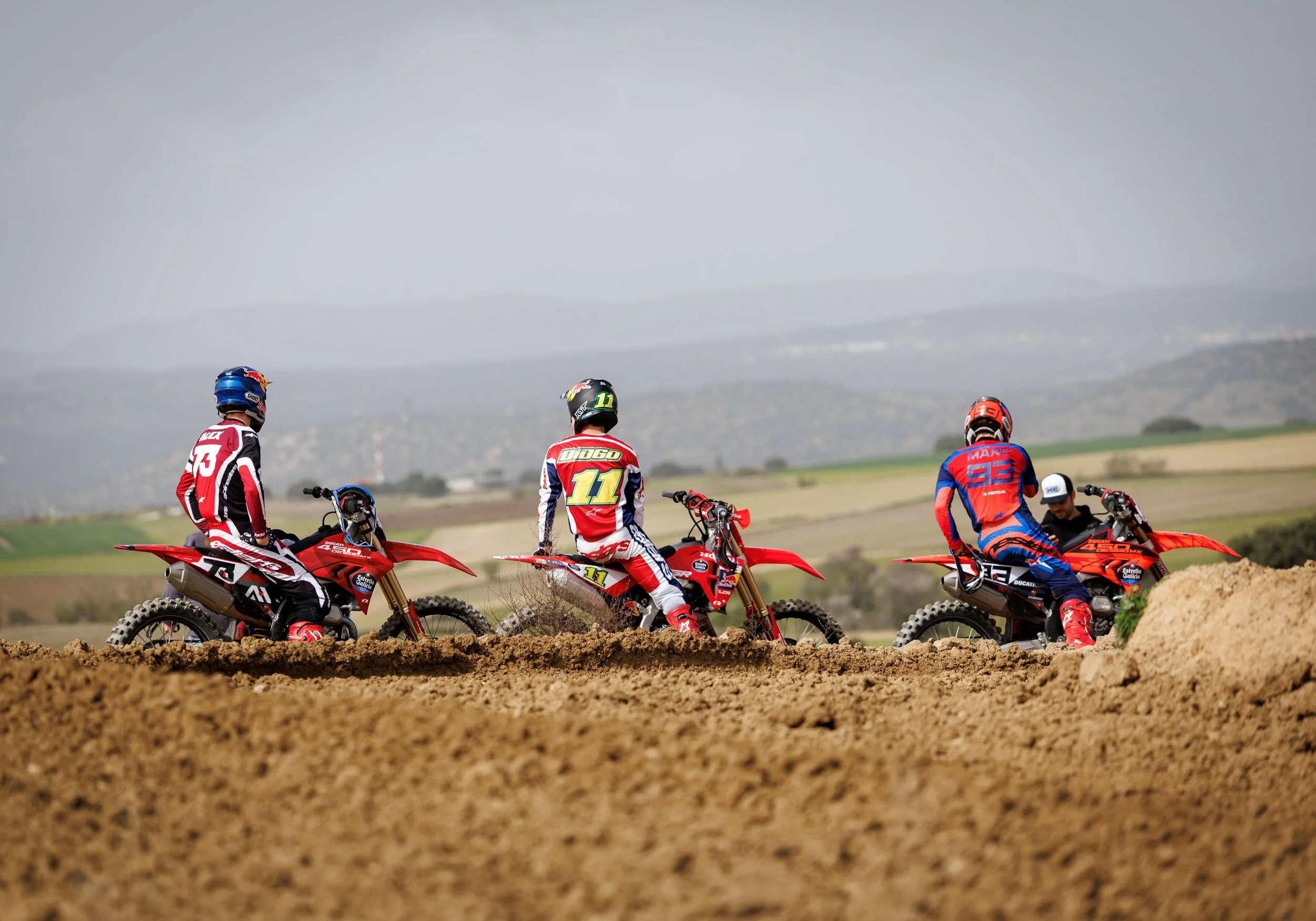 Cuatro pilotos de motocross en un campo, de espaldas, con un paisaje de colinas en el fondo