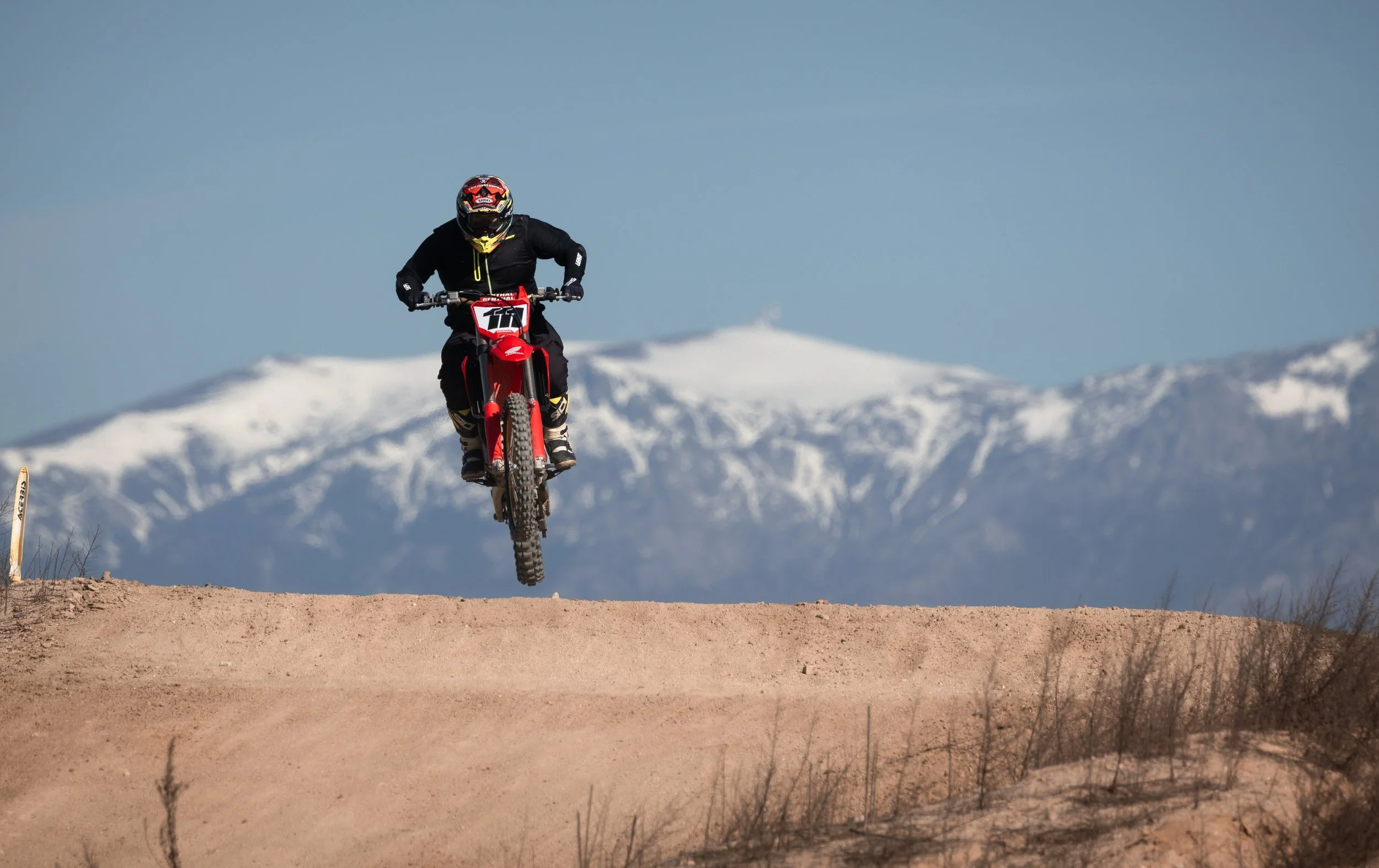 Persona en bicicleta de motocross saltando sobre un camino de tierra con montañas cubiertas de nieve al fondo.