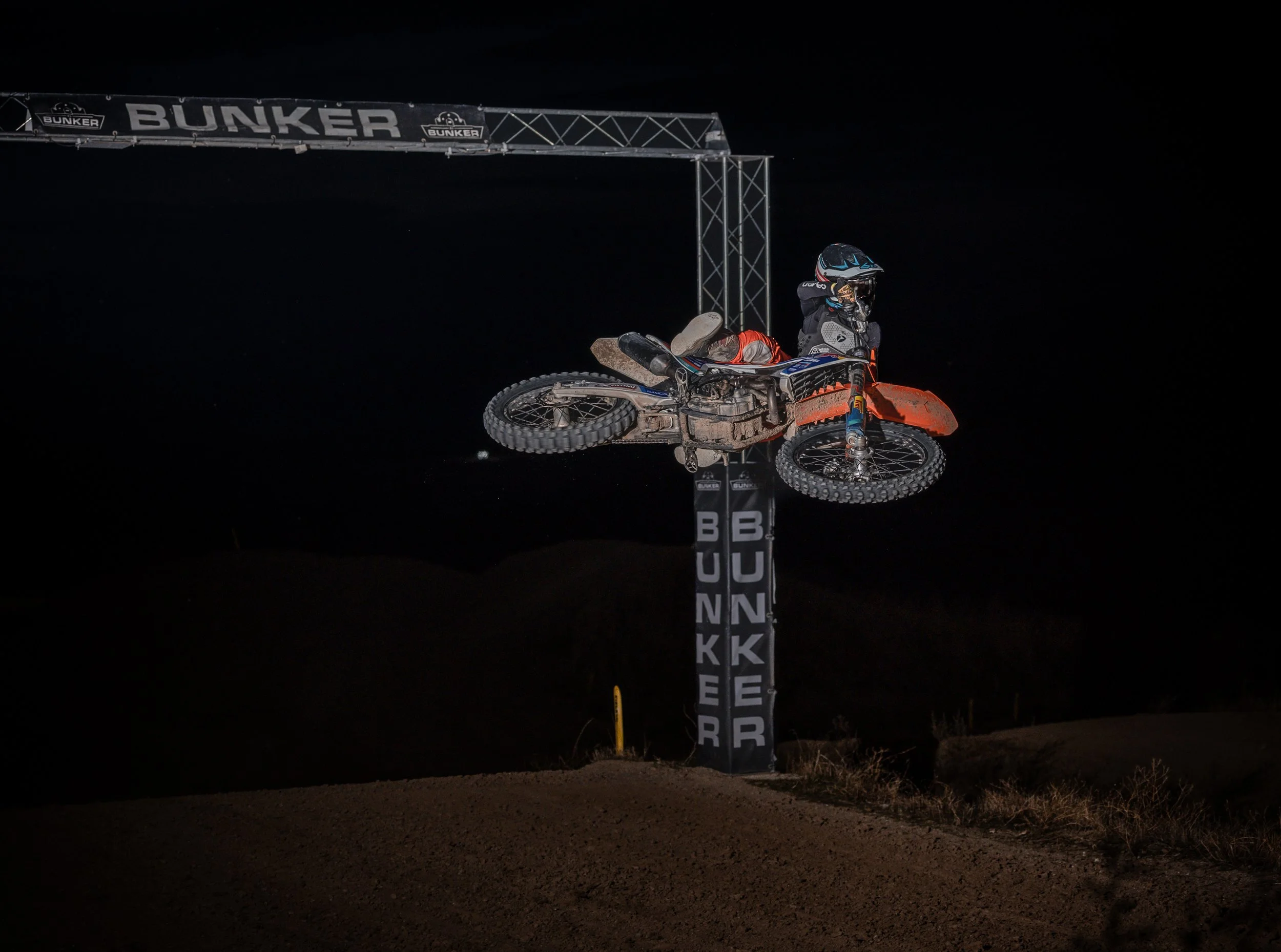 Un piloto de motocross realiza un truco en el aire en una competencia nocturna, con un arco que dice 'BUNKER' en el fondo.