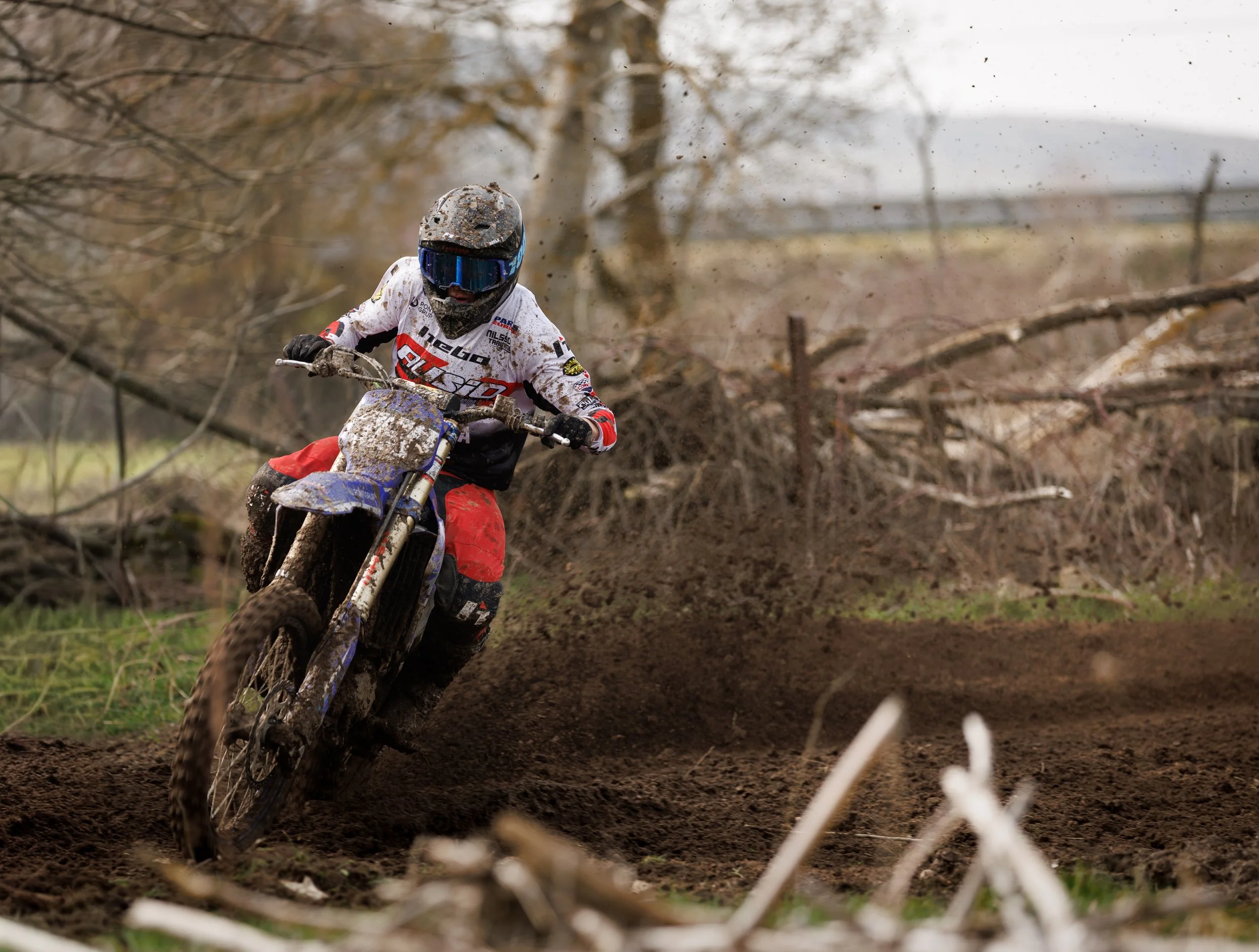 Una persona montando una moto de motocross en un terreno de tierra, con árboles y ramas caídas en el fondo.