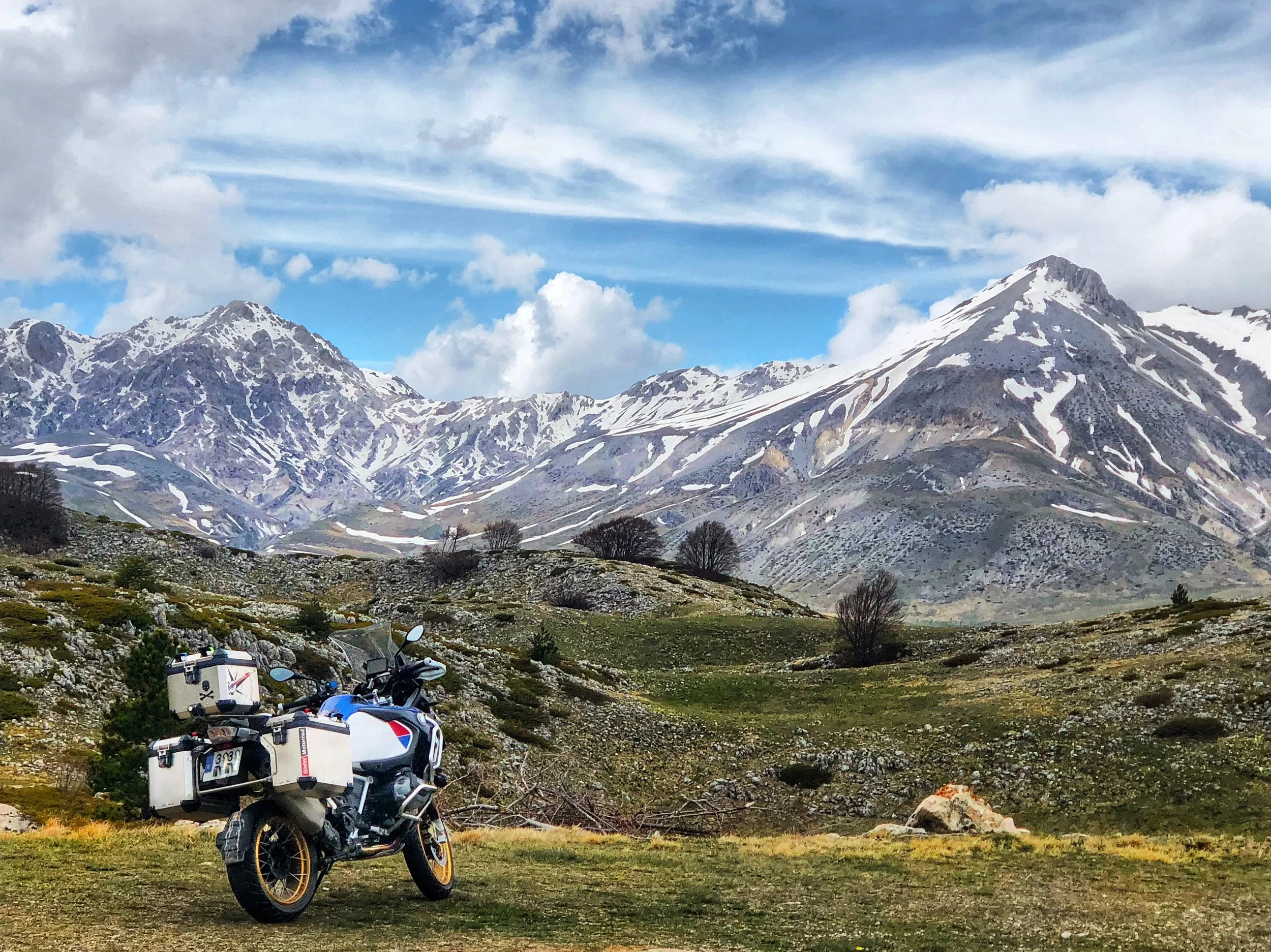 Una motocicleta en un paisaje montañoso con picos cubiertos de nieve, algunos árboles dispersos y un cielo nublado.